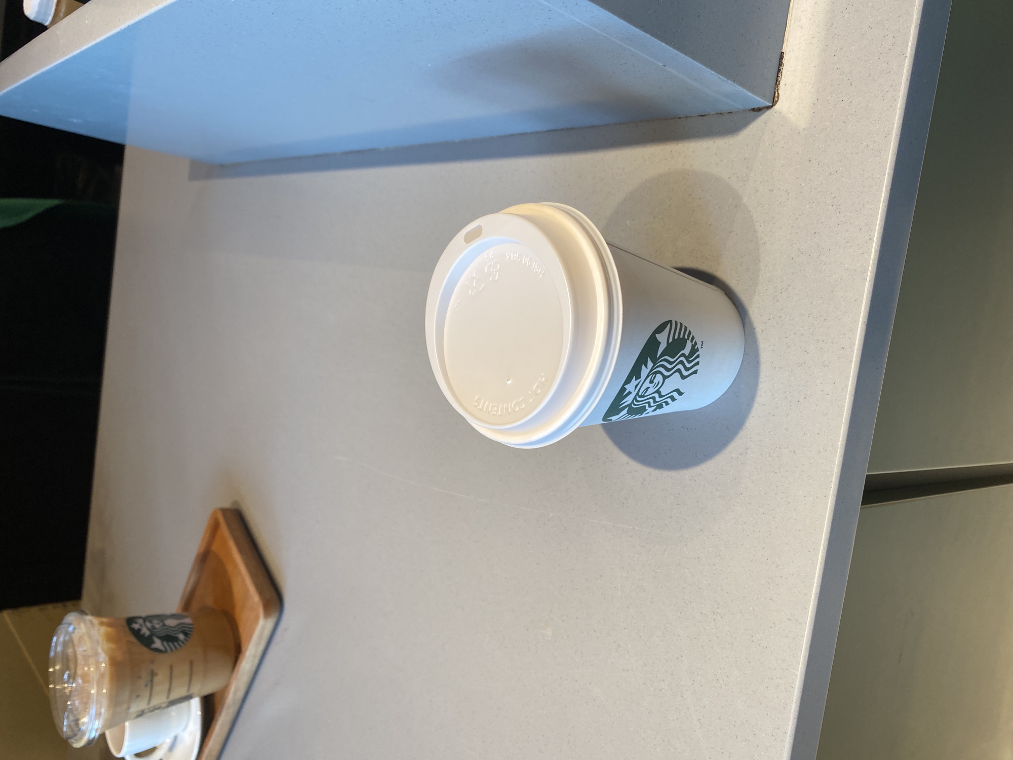 A high-angle, medium-shot depicts two Starbucks coffee cups on a light grey countertop. One cup is to the right, sealed with a white lid and emblazoned with the Starbucks logo in green. To the left and further away is another coffee cup, seemingly filled with coffee, sitting on a small, square, wooden tray. The background includes a shelf above the countertop and a blurry green and black area to the left. The location appears to be indoors, potentially in a Starbucks shop in Belgrade, Serbia.