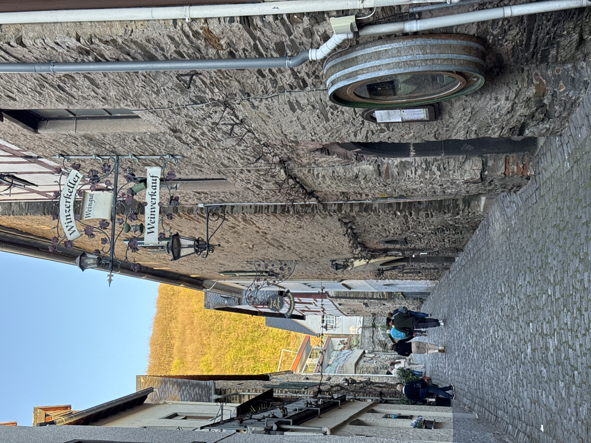 An eye-level, vertical shot depicts a narrow cobblestone street in Beilstein, Germany. The buildings on either side have stone walls, with some displaying signs. A sign with "Winzerkeller Weiningut" is visible. A decorative barrel ring is affixed to the wall. People are walking down the street away from the viewer. The street ascends toward an area with a view of hills. The sky is clear blue.