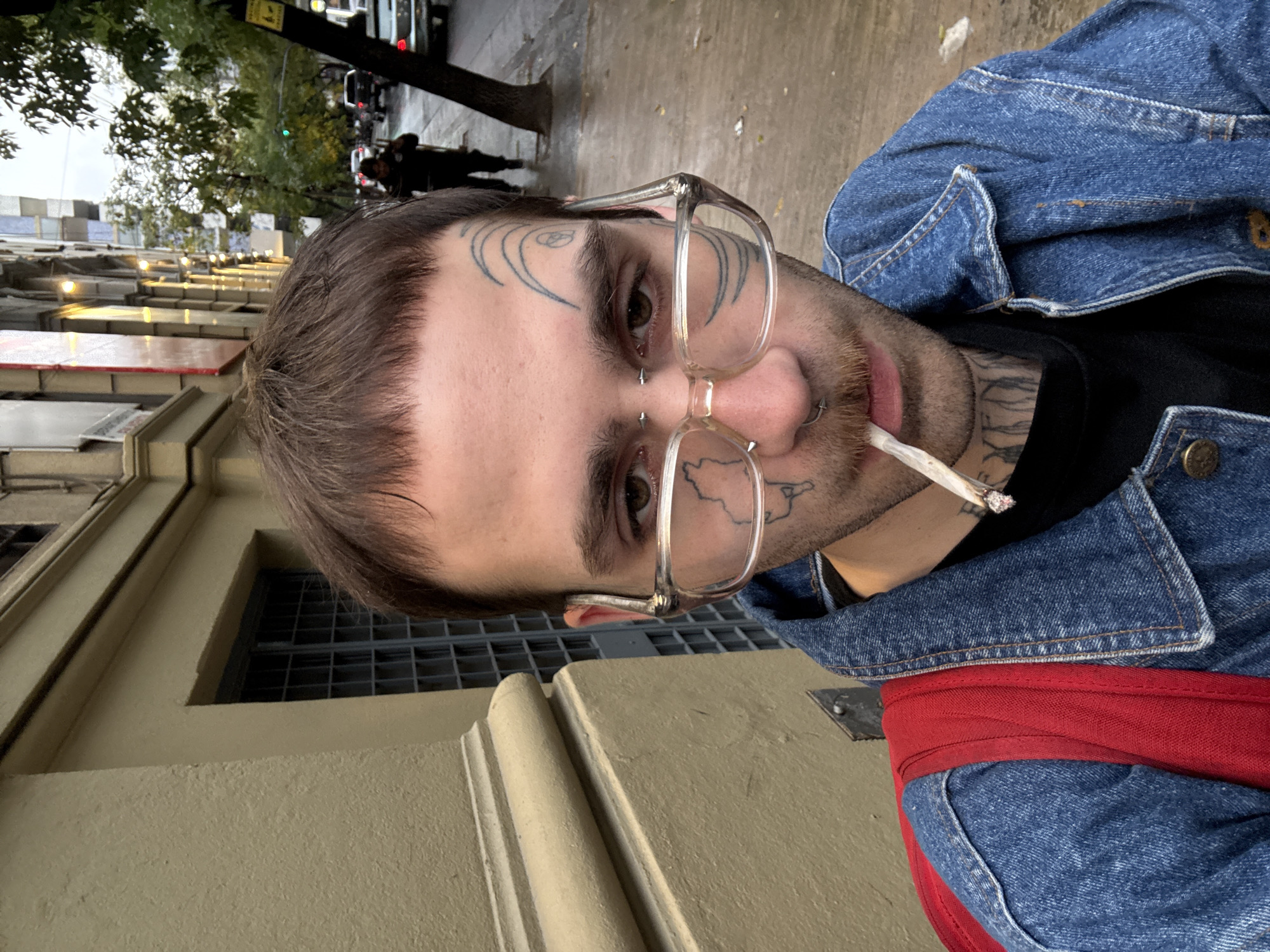 A man is standing on a sidewalk in Buenos Aires, Argentina. He is wearing a blue denim jacket, a black t-shirt, and clear-rimmed glasses. He has facial tattoos and piercings. He is holding a lit cigarette in his mouth. A red backpack is on his back. In the background, there is a building with barred windows and a street with cars and pedestrians.