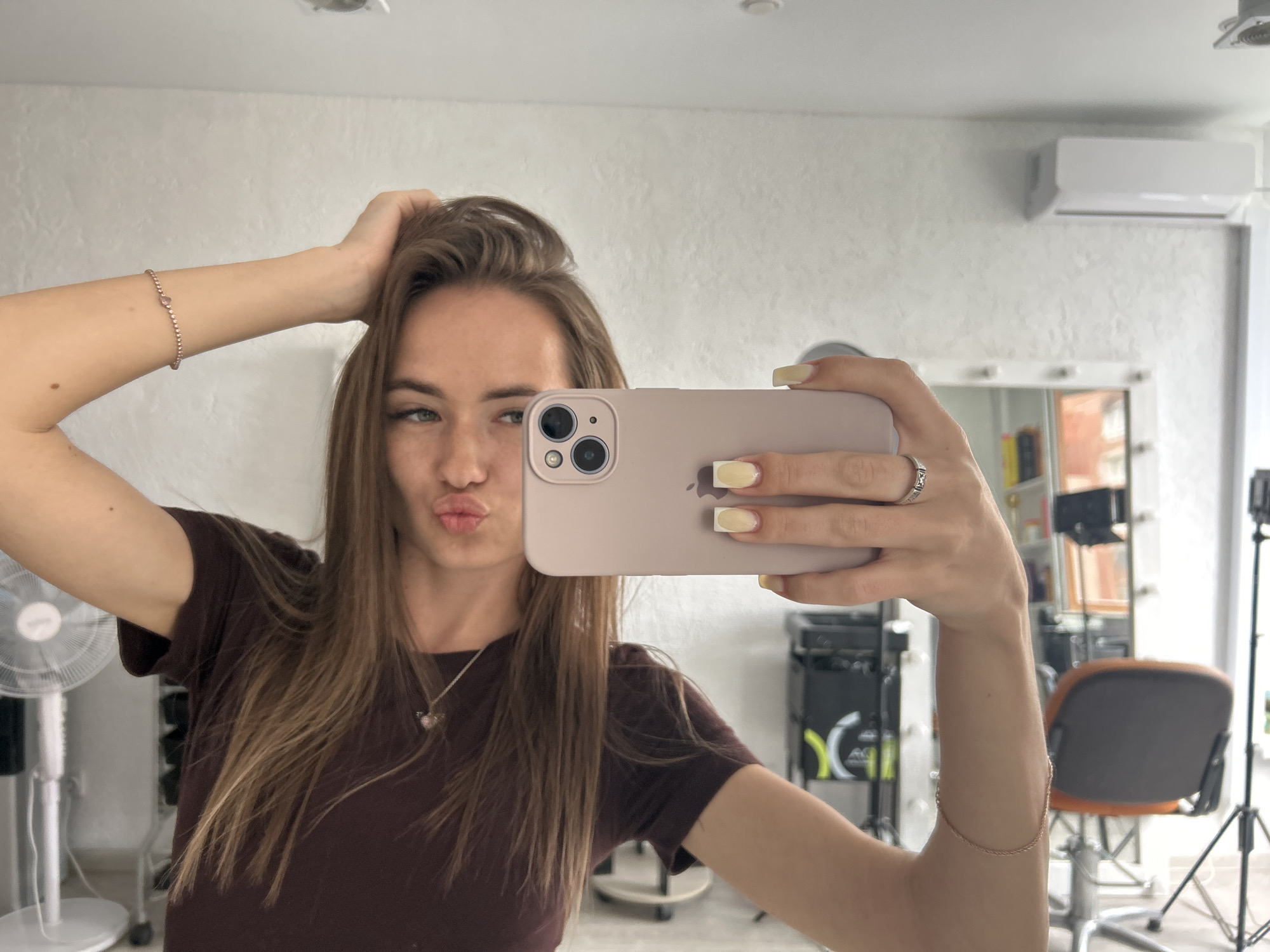 A young woman is taking a selfie in a brightly lit room. She has long, light brown hair, is wearing a dark maroon t-shirt, and has her left hand resting on her head. Her right arm is extended, holding a light pink smartphone, and she is puckering her lips. She is wearing a bracelet on her left wrist and a ring on her right ring finger. The room appears to be a salon or studio, with mirrors, hair styling equipment, and an air conditioning unit visible in the background. A white fan is also present to the left of the woman.