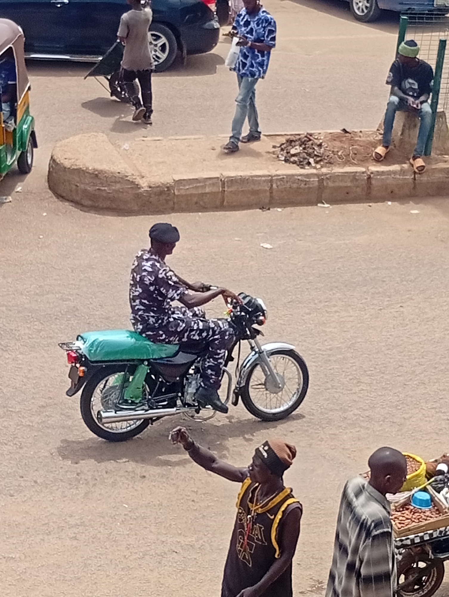 In Bukuru, Nigeria, a man wearing a black beret and a patterned dark and light uniform sits astride a motorcycle with a green-covered seat on a paved road. Another man, wearing a dark tank top with yellow trim and a beanie, stands nearby with his arm raised. A third man stands near a cart laden with peanuts and a blue container. In the background, a man in a blue patterned shirt walks on a raised curb, and another man sits on the curb edge near metal fencing. A green and brown tuk-tuk is parked on the side of the road. Another individual pushes a green wheelbarrow in the background near a dark-colored car.