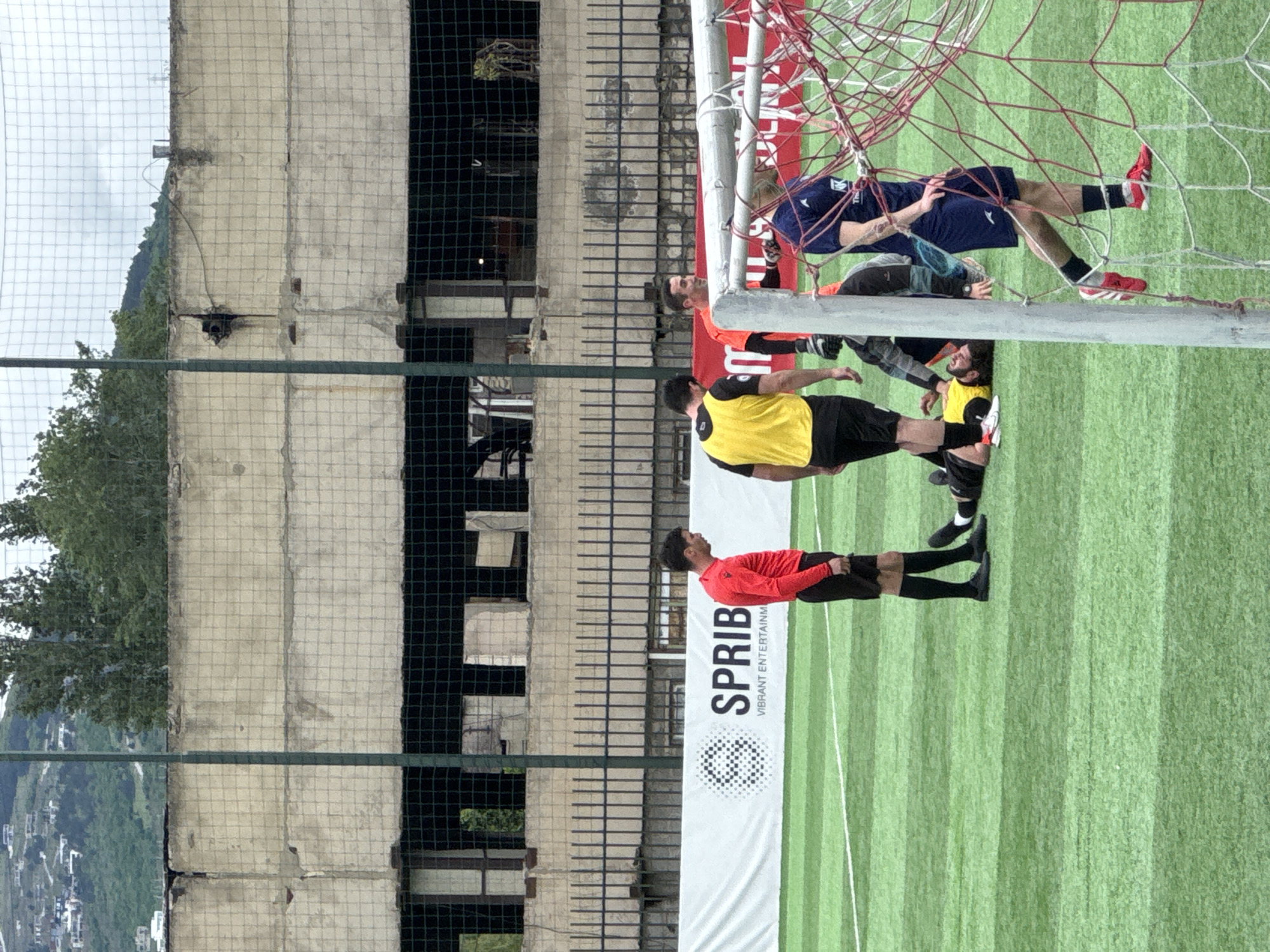 The image depicts a soccer match or practice in Tbilisi, Georgia. The backdrop includes a weathered, concrete building adjacent to the field. A soccer goal and netting are prominent in the foreground. Several soccer players are visible on the field, wearing various uniforms, including yellow, red, and blue. A white banner with the word "SPRIB" is present beside the field. The image includes sections of greenery and a view of a mountain range in the background.