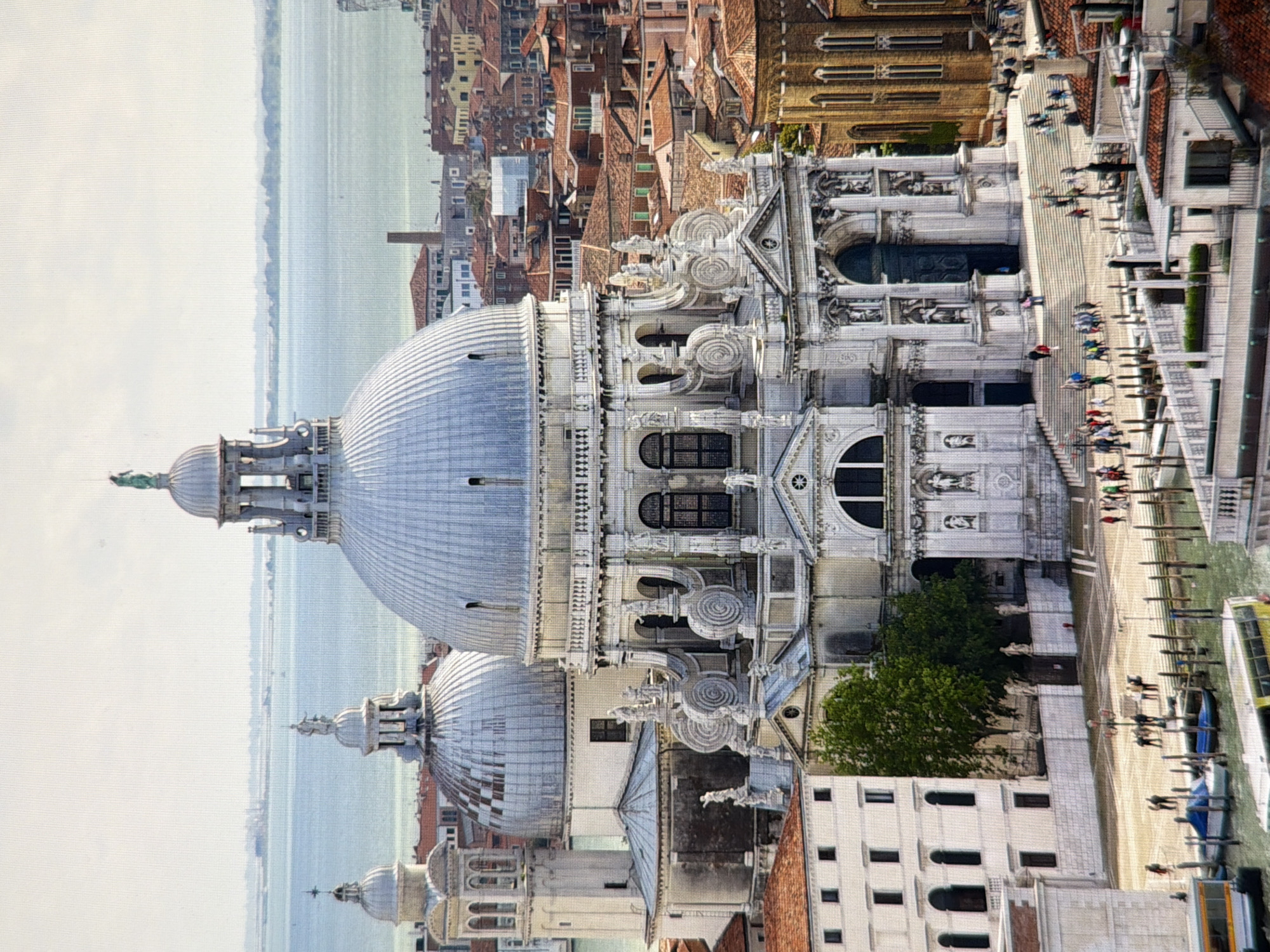 The image shows a wide shot of the Santa Maria della Salute church in Venice, Italy. The church is a prominent landmark with a large dome and a white facade. It is situated on the Grand Canal, with other buildings and the water visible in the background. The scene is bustling with activity, with many people walking around the church and boats on the water. The sky is overcast, and the lighting is soft, creating a serene atmosphere. The architecture is grand and ornate, characteristic of Venetian Baroque style. The church's dome dominates the skyline, flanked by smaller domes and bell towers. In the foreground, a canal with boats and people can be seen. The overall impression is one of historical significance and architectural beauty. The image does not depict Kyiv, Ukraine.