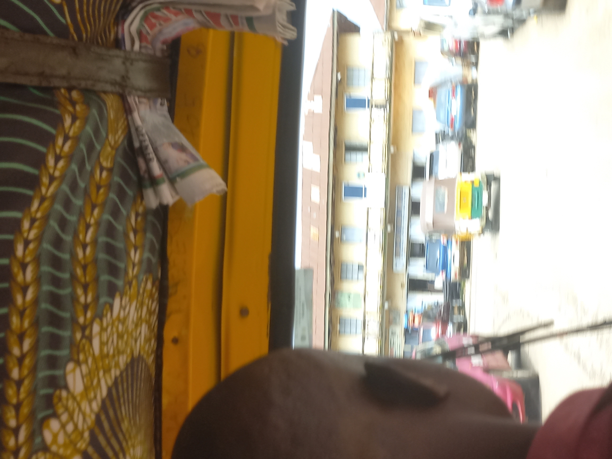 The image captures a street scene, possibly in Jos, Nigeria, taken from inside a yellow vehicle. In the foreground, the back of a person's head and shoulder are visible, suggesting the photographer is a passenger. The interior of the vehicle features a yellow canopy and a patterned fabric with brown, green, and yellow designs. Outside, buildings with light-colored facades and brown roofs line the street. Several vehicles are present, including a pink car, a blue car, a light-colored SUV, and a three-wheeled auto-rickshaw (Keke Napep) painted yellow and green. The overall impression is that of a typical urban environment.
