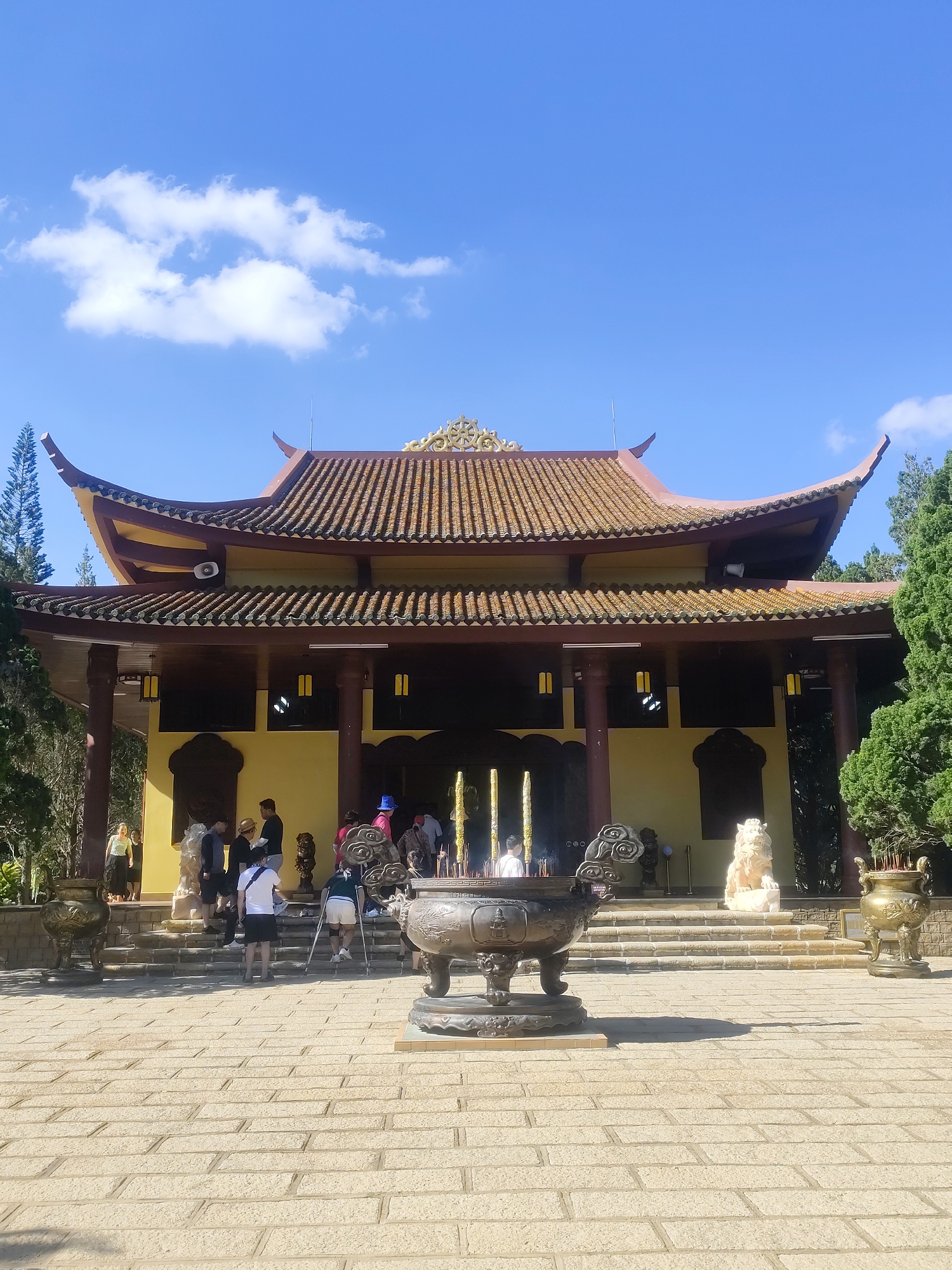 This is a medium-shot, eye-level, outdoor, daytime, vertical, full-color photograph taken from a paved courtyard looking towards a large, yellow Buddhist temple with a curved, tiled roof. The temple's roof is adorned with ornate golden decorations at its peaks. In front of the temple, there are several steps leading up to the entrance. Several people, appearing to be tourists, are gathered on the steps and in front of the temple. Some are looking at the temple, while others are engaged in conversation. In the foreground, a large, ornate, dark bronze incense burner sits on the paved courtyard, with smoke rising from it. Another similar incense burner is visible to the right, and a golden urn is positioned on a stone base to the far right. The courtyard is paved with light-colored rectangular stones. The background features lush green trees and a bright blue sky with scattered white clouds. The overall scene suggests a visit to a temple or a significant religious or cultural site. This appears to be the Truc Lam Zen Monastery, located on a hill overlooking Xuan Huong Lake in Da Lat, Vietnam.