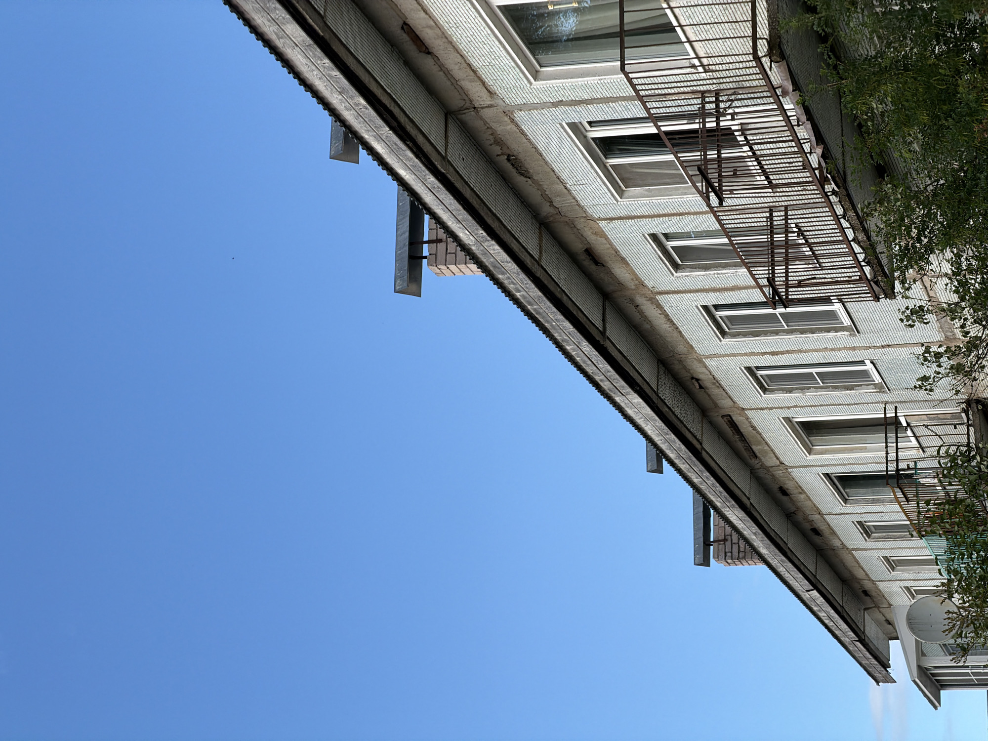 The image shows the facade of a multi-story building in Moshenskoe, Russia, under a clear blue sky. The building appears to be aged, with a tiled exterior and balconies with rusty railings. A sloping corrugated metal roof overhangs the top of the building. Chimneys with metal caps are visible on the roof. Green foliage is seen at the base of the building, obscuring the ground level. No people are visible in the image.