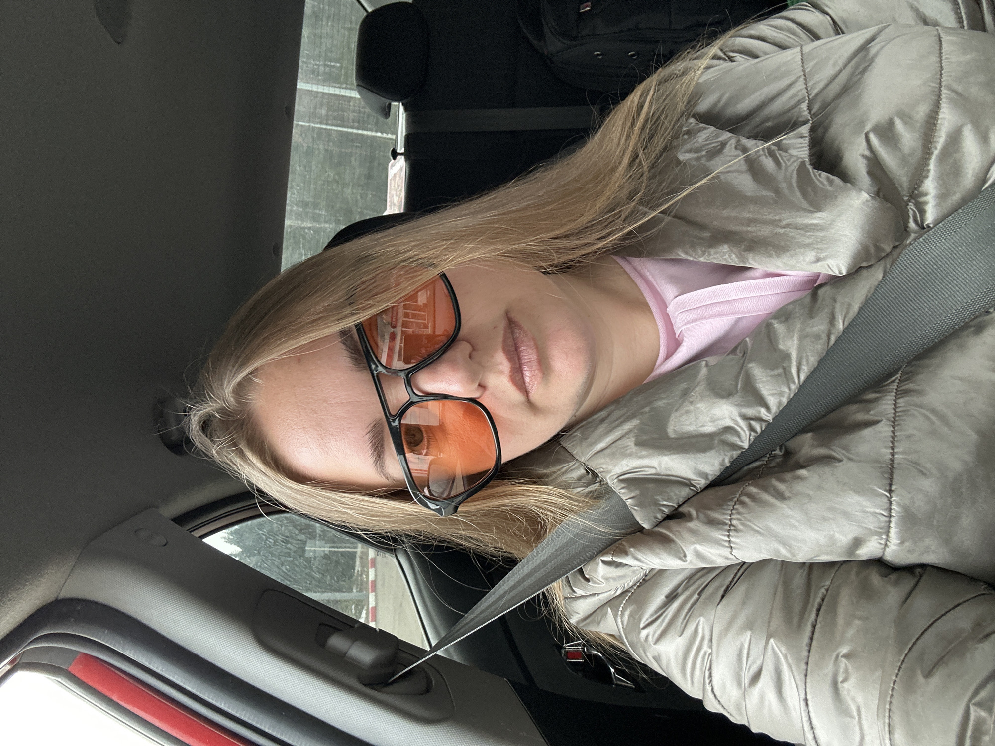 A fair-skinned woman with blonde hair is sitting in a car, wearing a silver puffer jacket, a pink shirt, and black-framed glasses with orange lenses. She is wearing a seatbelt. The car appears to be in Chaikovsky, Russia, based on the building reflected in her glasses. Other items in the vehicle include the car's interior and an unidentified bag in the back.