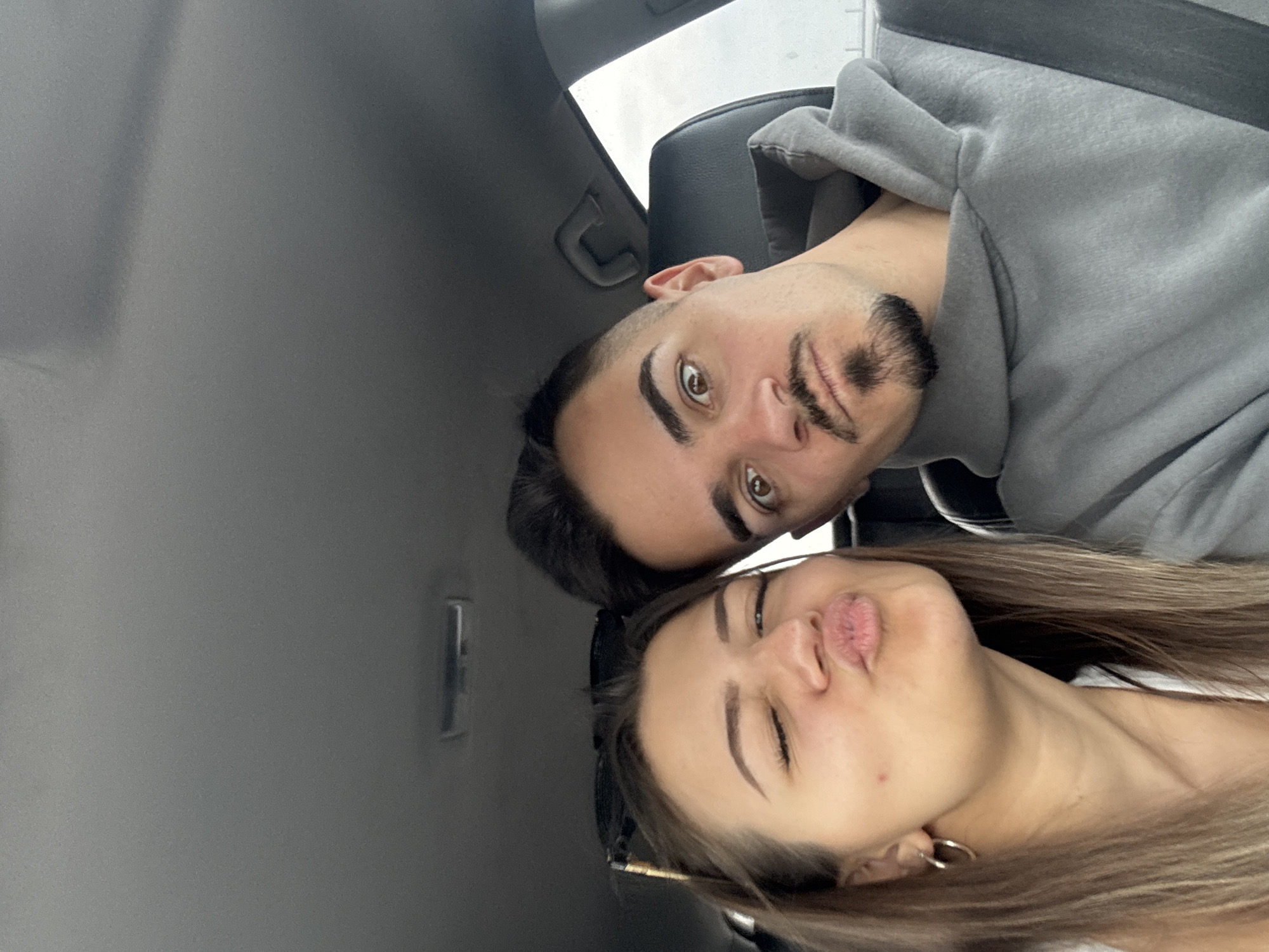 Two individuals are visible in the image, positioned within the interior of a vehicle. A male figure, sporting a beard and wearing a gray hoodie, is located towards the upper portion of the frame. A female figure with long, light brown hair, is situated below him, pursing her lips into a pouting expression. The interior of the car is visible, with gray panels and black seats. The location in Cherna Gora, Bulgaria, is not discernible from the image.