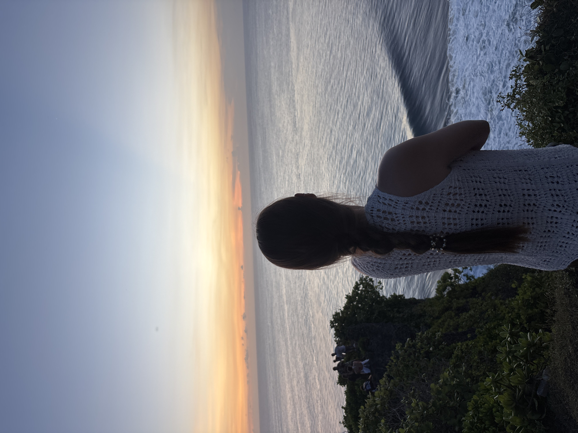 A sideways, eye-level shot shows a woman looking out at the ocean at sunset in Kuta Selatan, Indonesia. She has dark brown hair pulled back in a braid. She is wearing a white, crocheted top. To her right, green foliage covers the cliffside and a group of people stand observing the view. The ocean is in front of her, and the sky at the horizon is yellow, fading into a light blue overhead. There is a wave in the ocean near the cliffside.