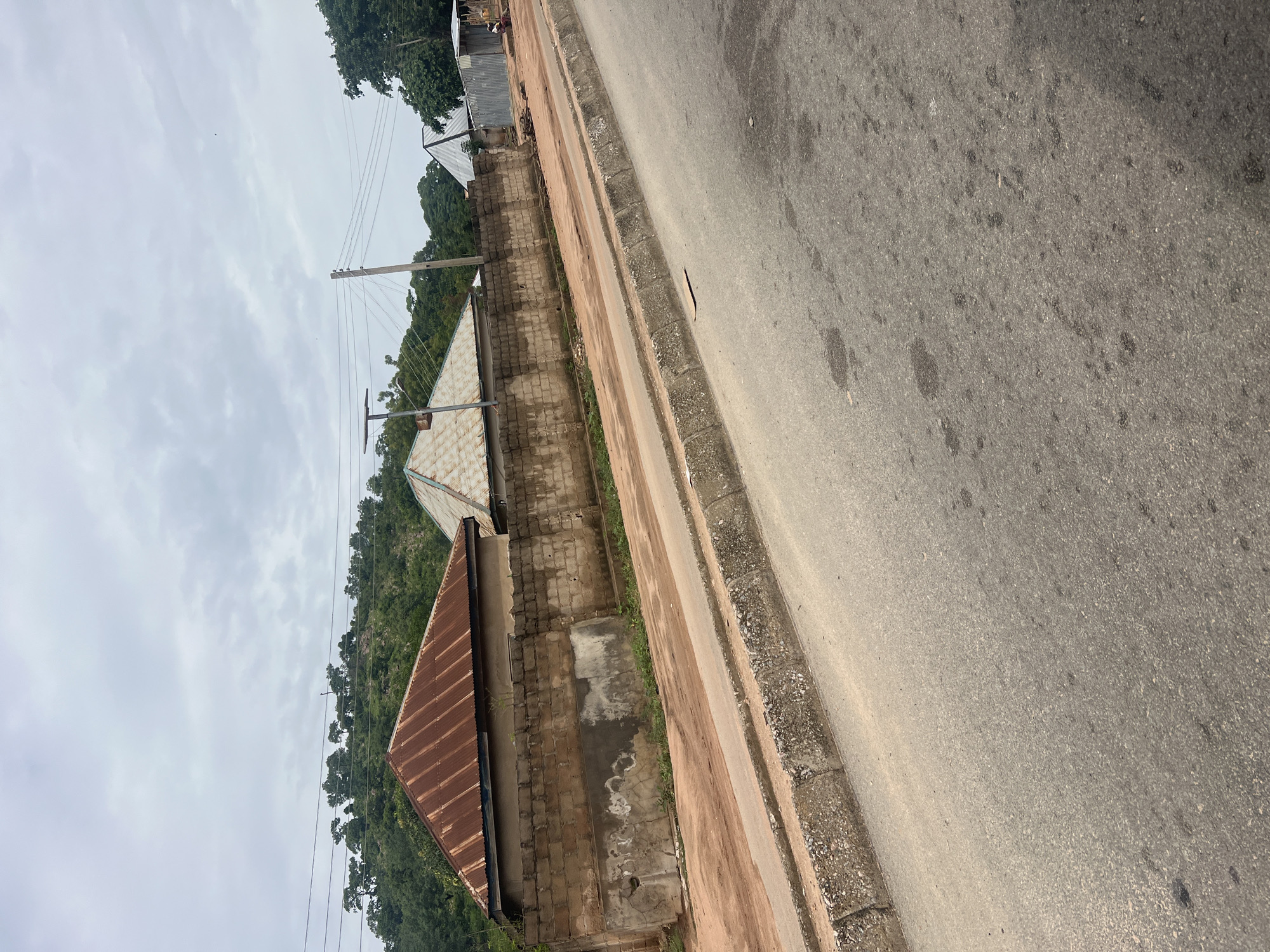 The image depicts a street scene in Minda, Nigeria, with a focus on a row of buildings behind a low, brick wall. The buildings have distinct corrugated metal roofs, one prominently rusted red and another lighter colored with diamond patterns. In the background, a lush, green hillside rises under an overcast sky. A paved road with a concrete curb occupies the foreground, suggesting it is a well-trafficked area. The overall impression is one of a semi-rural or developing urban environment.