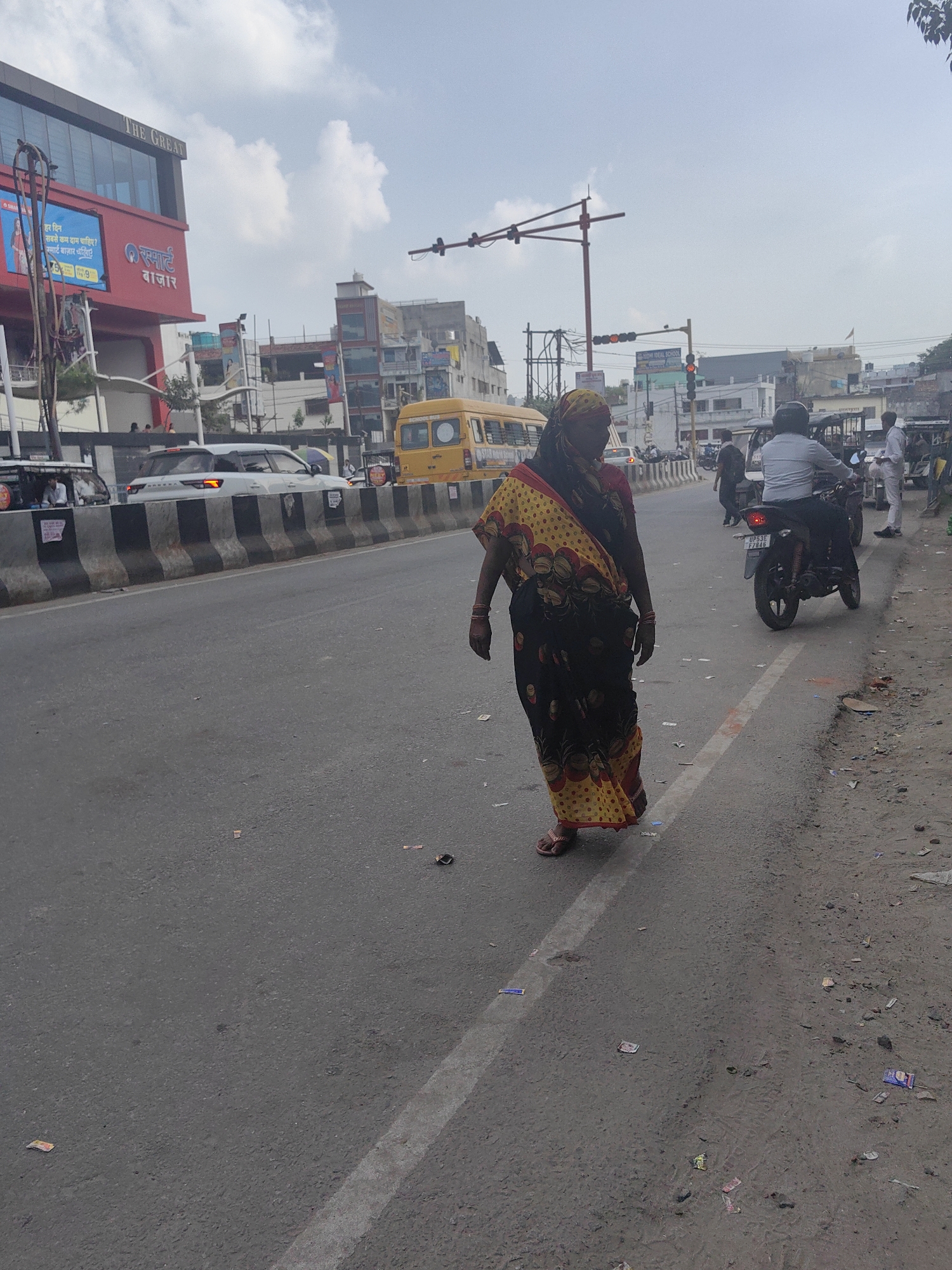 An Indian woman in a yellow and red sari walks on the side of a busy street in Gorakhpur, India. She is facing away from the camera, and appears to be mid-stride. The street is lined with concrete barriers, and there are several vehicles visible, including a white SUV, a yellow school bus, and a motorcycle with a rider. Buildings line the street, some of which appear to be shops or businesses. The sky is cloudy, and the overall lighting suggests it is daytime. The ground is littered with small pieces of paper and debris.