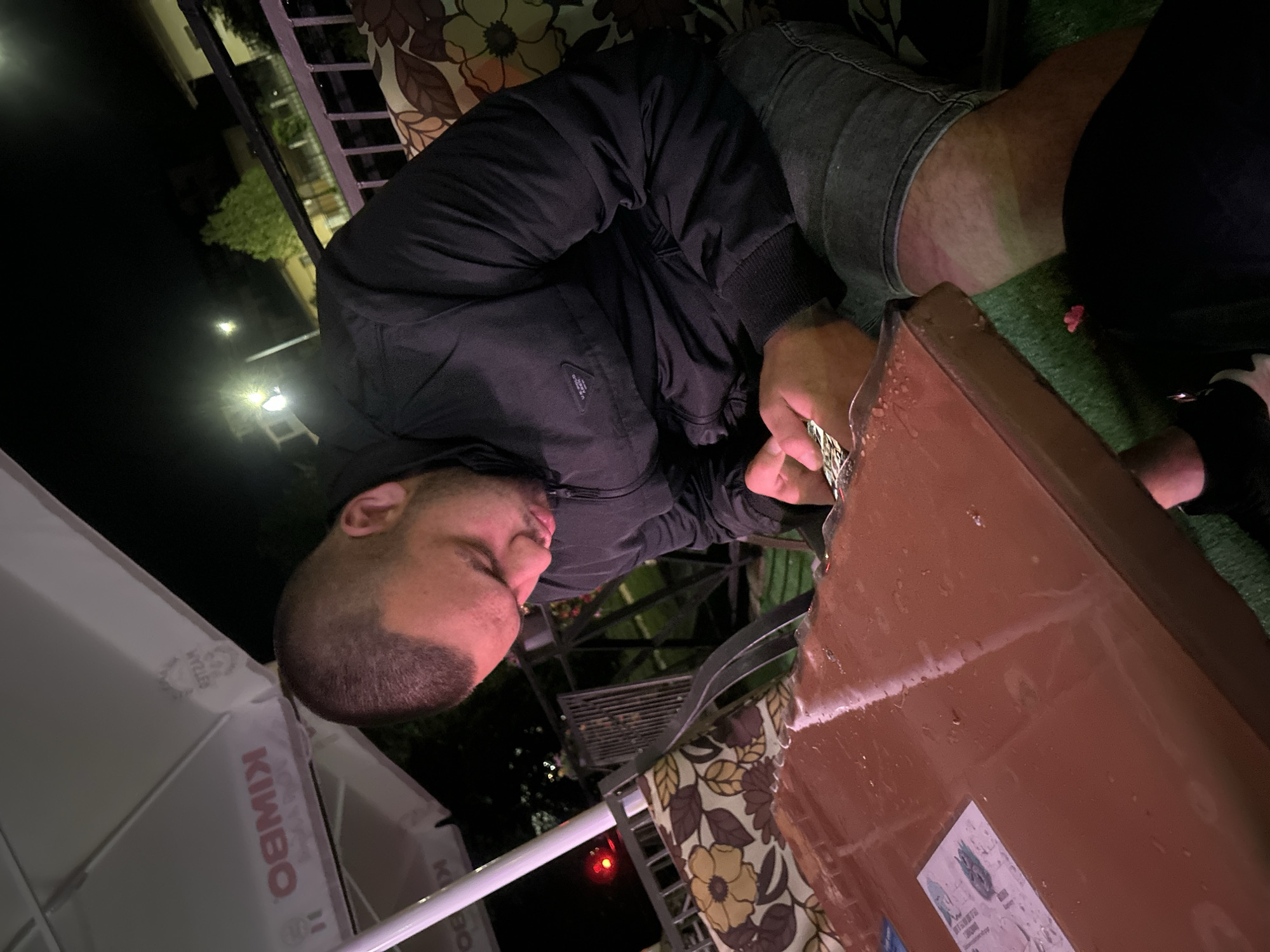 The image depicts an outdoor night scene, likely a patio or café area in Bania, Bulgaria. The primary subject is a man, viewed from the side, sitting at a brown table. He has short dark hair and a beard/stubble, and is wearing a dark, possibly black, long-sleeved jacket and denim shorts. His hands are near the edge of the table, which appears wet, possibly from condensation or a spill.
The setting is dark, illuminated by artificial lights, including distant streetlights visible in the background. To the right of the man, another person's lower body is partially visible, wearing what appears to be part of a black and white garment or shoe. The ground beneath the table is covered in artificial green grass, with a small pink object, possibly a flower petal, resting on it.
Other objects in the scene include a patterned cushion with a floral design in brown, yellow, and green, suggesting outdoor furniture. A large white umbrella is visible on the left, displaying the brand names "KIMBO" and "KRYZAN" vertically. A sticker with some unreadable text is affixed to the wet brown table. In the mid-ground, beyond the man, other dark outdoor furniture and a metal railing are faintly visible, further confirming an outdoor commercial or recreational space. The overall atmosphere is relaxed, typical of an evening gathering.