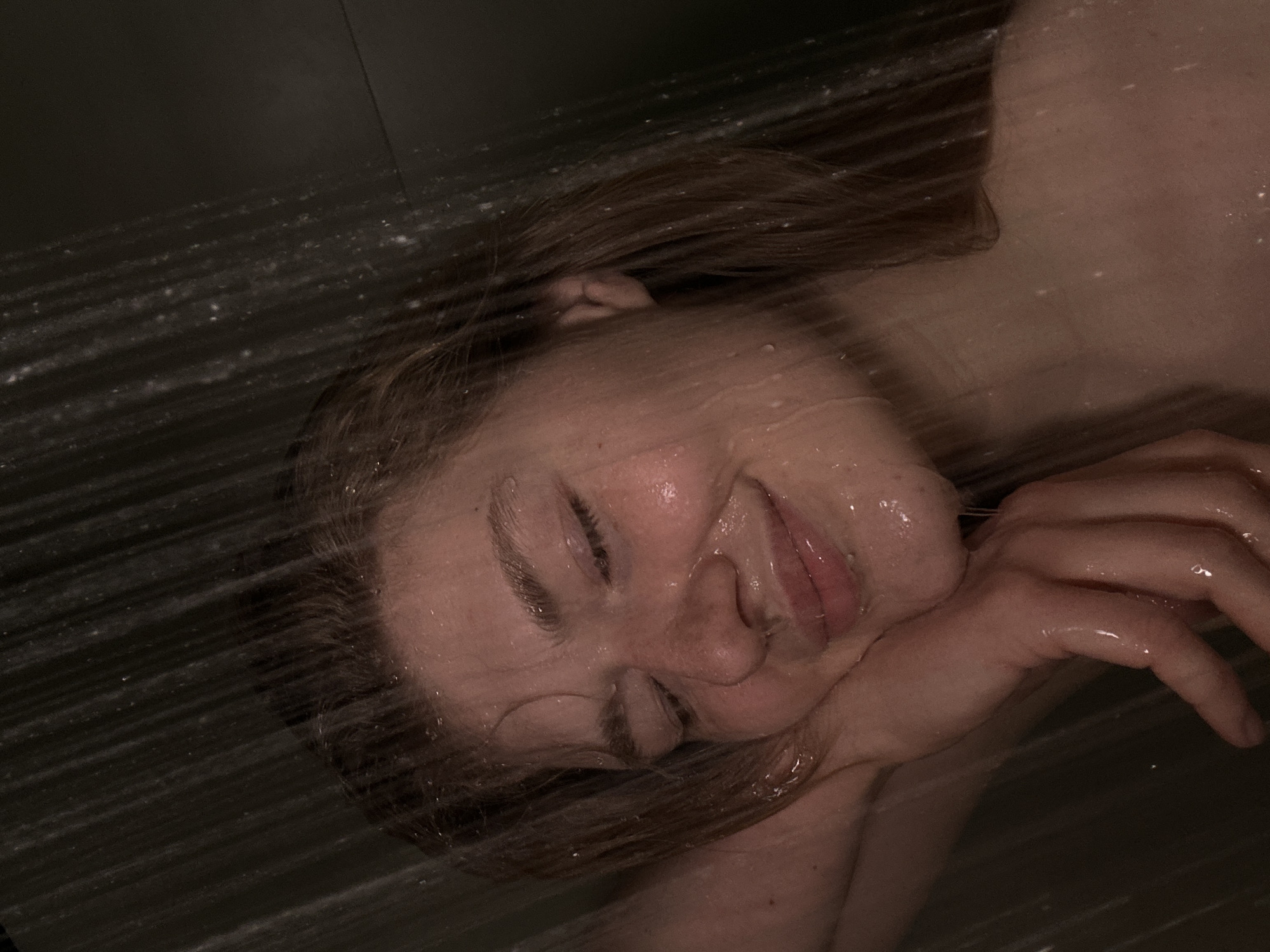 A person is shown lying down in what appears to be a shower, with water running over their face and body. The person has fair skin and light brown hair. Their eyes are closed, and they are smiling slightly. Their head rests on their hand. The environment is dark, and the focus is on the person and the water. There is no indication of the city of Kazan, Russia.