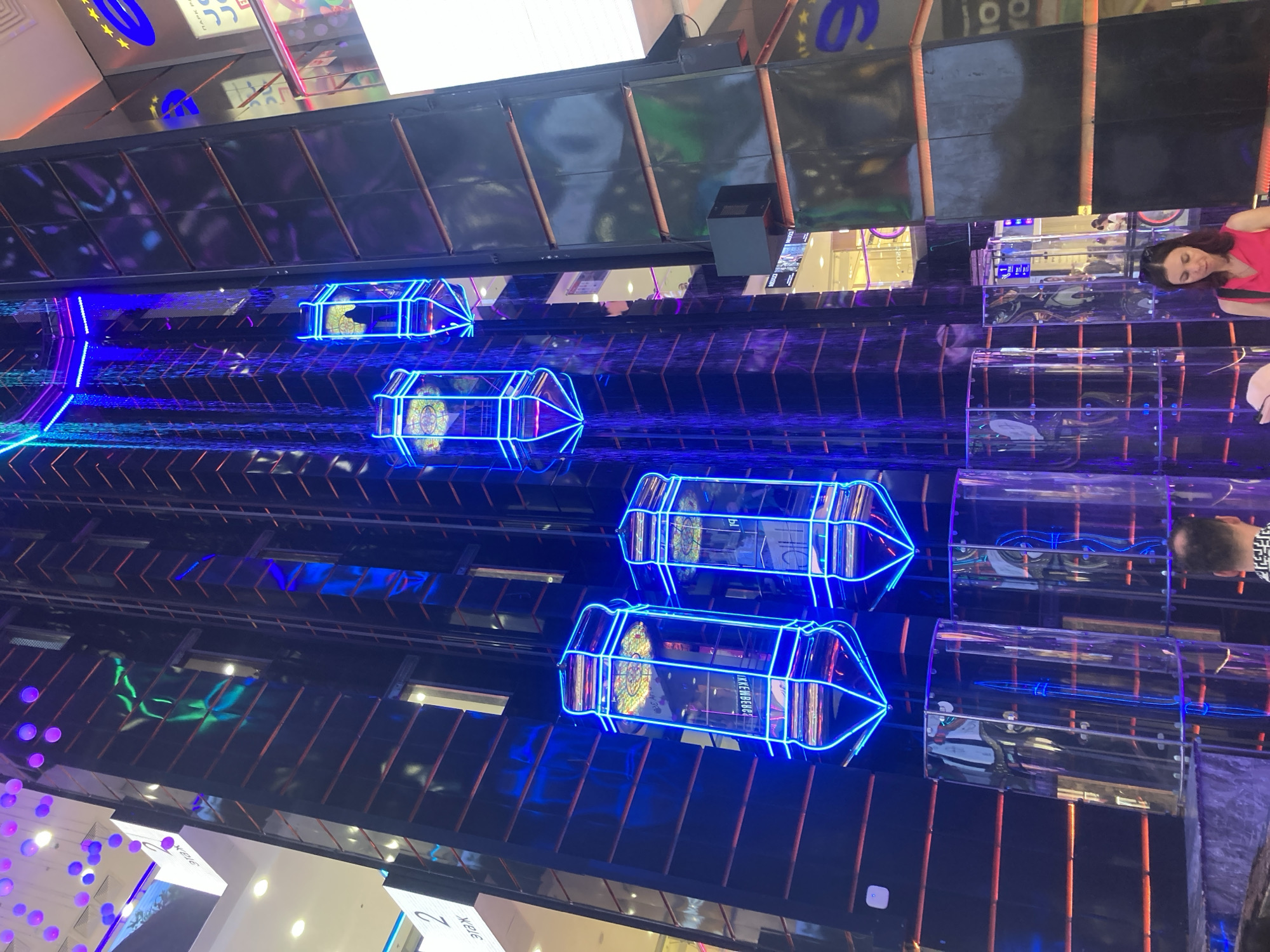 The image captures the multi-story central atrium of Afimall City shopping center in Moscow, Russia. Six glass-walled, panoramic elevators, each illuminated with bright blue neon outlines and featuring a faceted, capsule-like design, are suspended within a tall, dark-paneled central structure. A prominent indoor waterfall, lit by blue and purple lights, cascades down the central structure adjacent to the elevators. The dark panels of the building facade are interspersed with horizontal reddish-orange accent lines. Signs displaying "2 ЭТАЖ" (2nd Floor) are visible on the left. In the foreground, a woman with dark hair, wearing a pink top, is shown from the chest up, looking downwards. Behind her, the back of a man's head, partially bald with dark hair, and wearing a patterned shirt, is visible. Reflections of interior lights and people are apparent on the reflective surfaces of the elevators and the surrounding building. Transparent cylindrical structures are positioned at the base of the central elevator shaft. A large blue 'e' logo adorned with yellow stars is visible on a dark panel on the right side of the image.