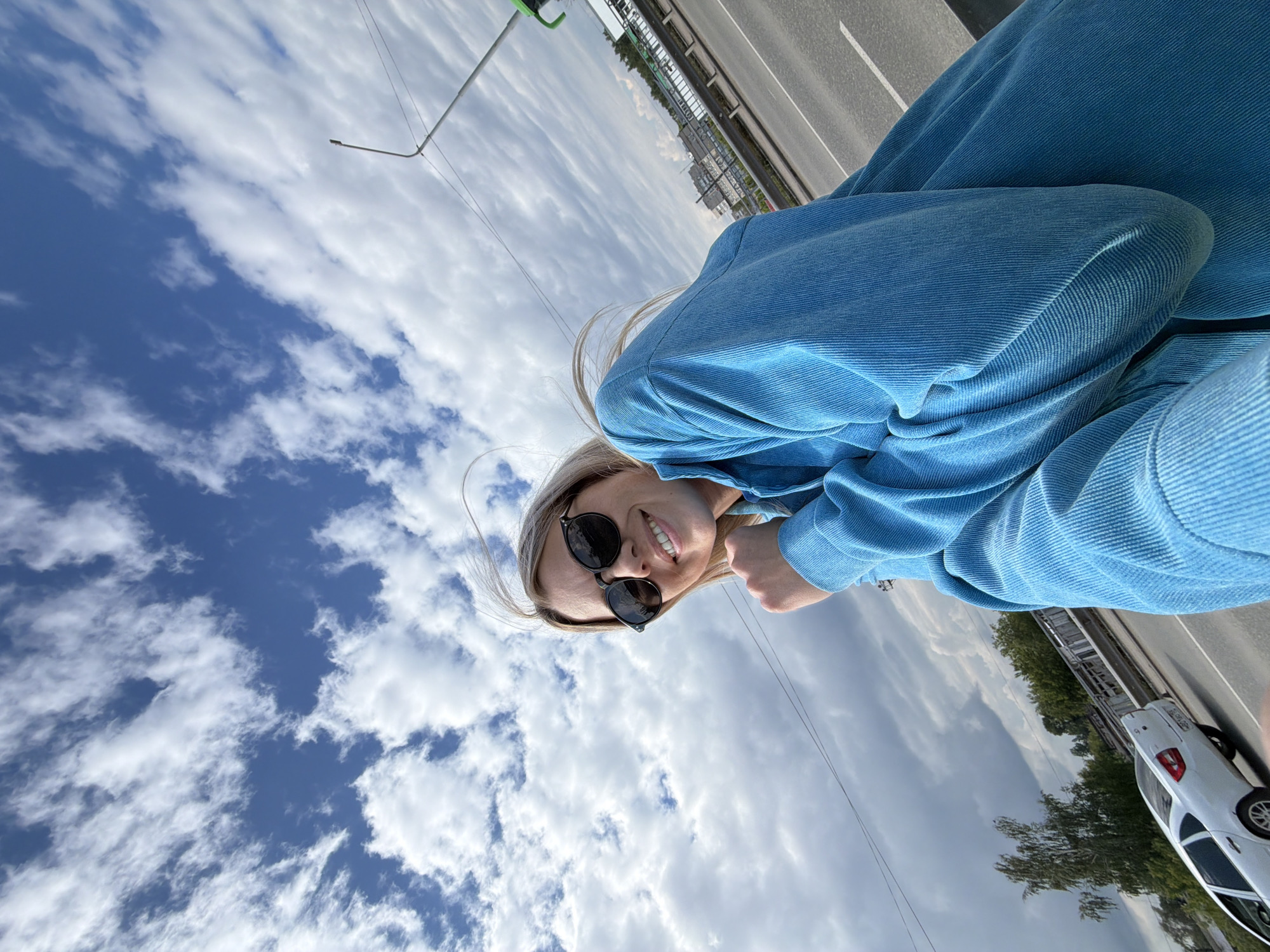 A bright portrait-oriented image captures a smiling young woman against a backdrop of a partly cloudy sky and a highway. The woman, with blonde hair and wearing black sunglasses, is positioned in the upper right of the frame, looking up at the camera with a wide smile. She is dressed in a vibrant blue, textured long-sleeved top, possibly corduroy or ribbed knit.
The sky, occupying a large portion of the left and upper parts of the image, is a clear blue with scattered white, fluffy cumulus clouds. To the right, a multi-lane highway or road extends into the distance, bordered by a metal guardrail. A white sedan car is partially visible on the far right of the road. Trees and some distant buildings or structures can be seen beyond the road, suggesting an urban or suburban setting. On the far left, a tall, slender pole, likely a street light, extends upwards.
The image appears to be a daytime selfie, characterized by bright, natural lighting. The woman's expression conveys happiness and a relaxed mood. The location is Chaikovsky, Russia. There is no visible text in the image.