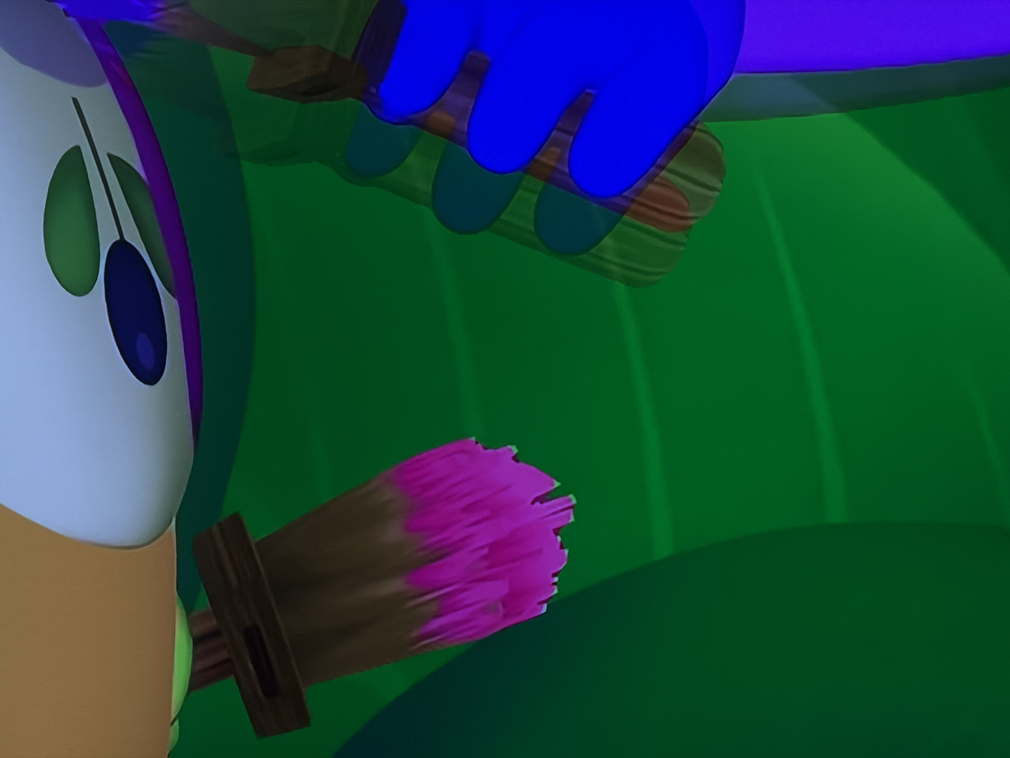 An animated scene depicts two cartoon characters engaged in painting. In the foreground on the left, a character with a light beige body and a white head is visible. It has large, oval, dark blue eyes and two small, green, leaf-like protrusions on either side of its face, topped by a purple band. This character holds a paintbrush with a brown wooden handle and pink bristles, loaded with pink paint. Above and to the right, a blue hand of another character holds a second paintbrush, also with a wooden handle, suggesting it is actively painting. The background consists of various shades of green, resembling foliage or a stylized natural environment, with undulating shapes and shadows. The overall aesthetic is brightly colored and typical of children's animation. There is no visible text, and specific time of day or weather cannot be determined from the stylized setting. The location context provided is Sofiivska Borshchahivka, Ukraine, though no direct visual cues within the image indicate this real-world location.