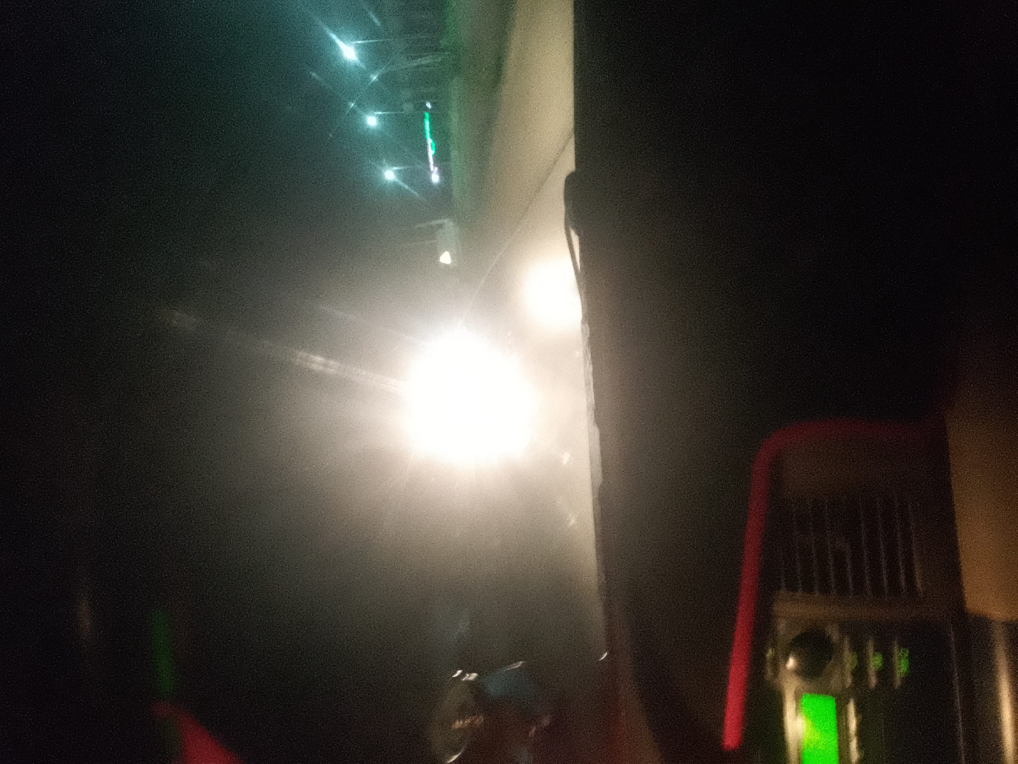 The image depicts a nighttime scene, likely from inside a moving vehicle, looking towards the right side of the frame. In the foreground on the right, a portion of a vehicle's interior is visible, including what appears to be a red-colored metal frame and a ventilation grille. The background is dominated by a bright, diffused light source, likely headlights from an oncoming vehicle, creating a hazy and washed-out effect.
Above and to the left of the main light source, several smaller lights are visible, arranged in a line and suggesting streetlights or lights from buildings. The overall environment appears to be a road or an urban area at night, with a sense of motion due to the blurred lights and the perspective. Given the context of Jalingo, Nigeria, this could be a scene on one of the city's major roads or a street in a populated area during nighttime hours. The image quality is grainy, typical of low-light photography. There are no discernible people in the image. The focus is primarily on the light sources and the immediate interior of the vehicle.