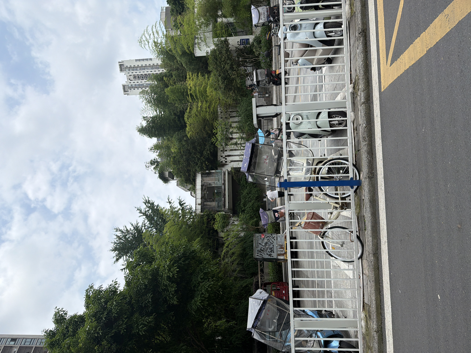 The image depicts a street scene in Chongqing High-tech Zone, China. A white metal fence separates the sidewalk from the roadway, where a yellow painted line is visible. Behind the fence, trees and foliage dominate the landscape, obscuring buildings. Parked scooters, bikes, and other vehicles are adjacent to the fence. A person wearing a white shirt is seen standing behind the fence.