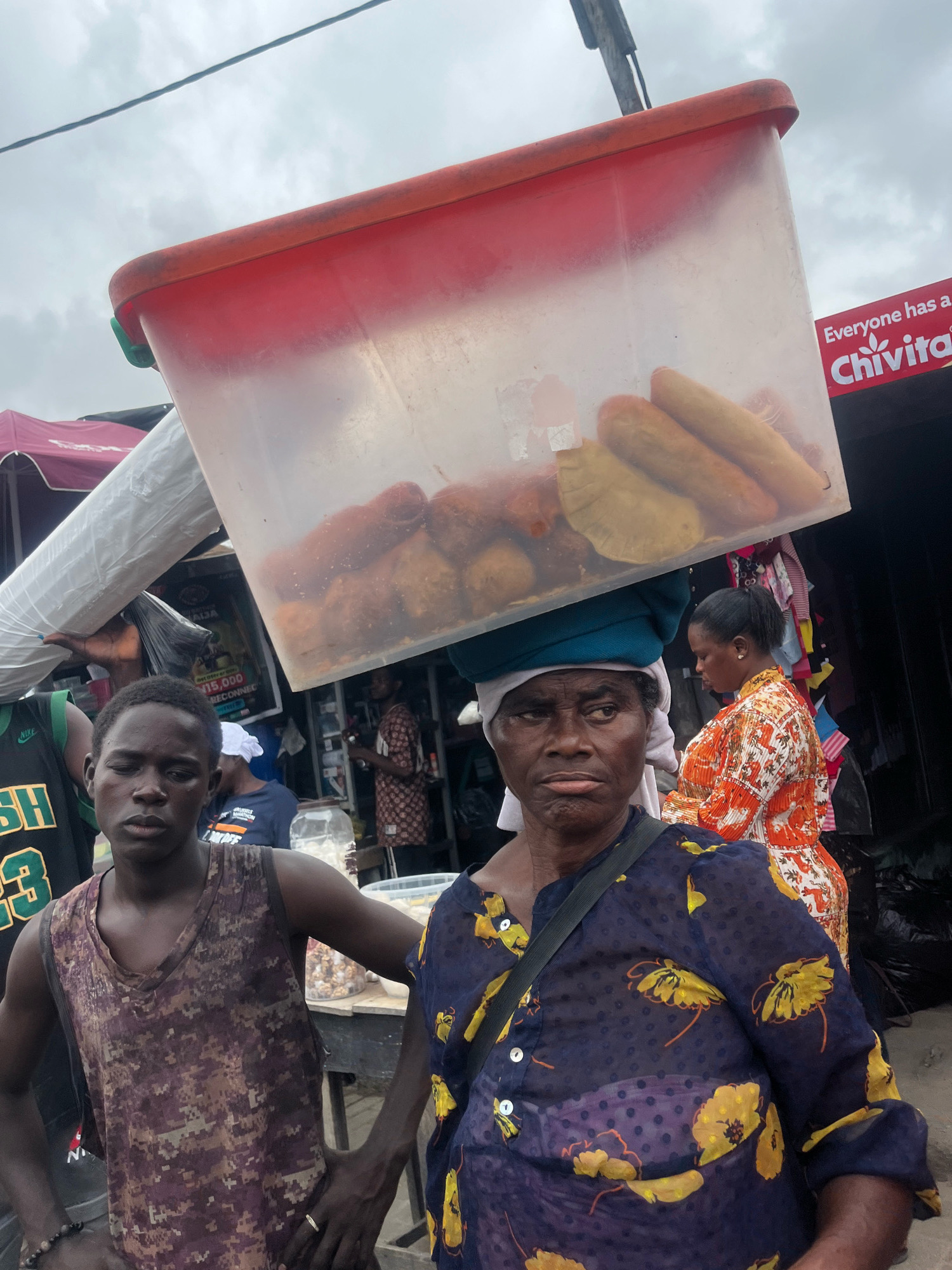 A medium shot captures a street scene in Lekki Phase I, Nigeria, under an overcast sky. In the foreground, a woman with dark skin, wearing a blue headscarf and a transparent dark blue top with yellow floral patterns, balances a large, clear plastic container filled with fried foods on her head. Her expression is serious, and she is looking slightly to her left. To her left, a young man with dark skin, wearing a camouflage-patterned tank top, stands with his hands at his hips, looking directly at the camera with a neutral expression. Behind them, a woman in an orange and white patterned dress is visible, as well as other market stalls and people. The overall scene depicts a bustling market environment.