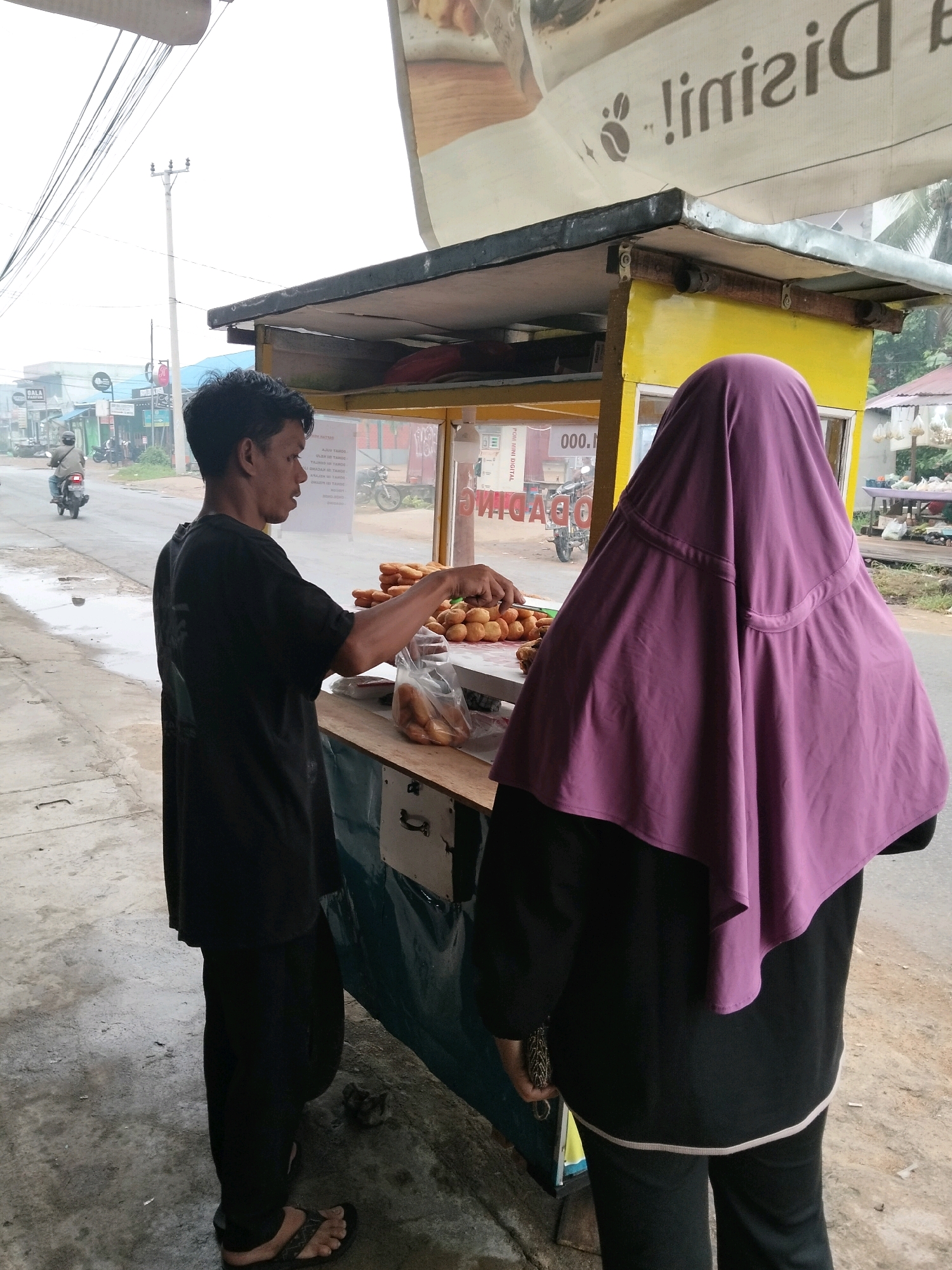 In Arang Limbung, Indonesia, a street vendor, a man wearing a black t-shirt and black pants, is selling fried pastries from a yellow and black food cart. He is interacting with a customer, a woman veiled in a purple hijab and wearing black clothing. The vendor is holding a plastic bag filled with pastries, and the woman is standing in front of the cart, facing away from the camera. In the background, a street is visible with a motorcycle rider passing by, and various buildings and utility poles line the roadside. The scene appears to be during the daytime.
