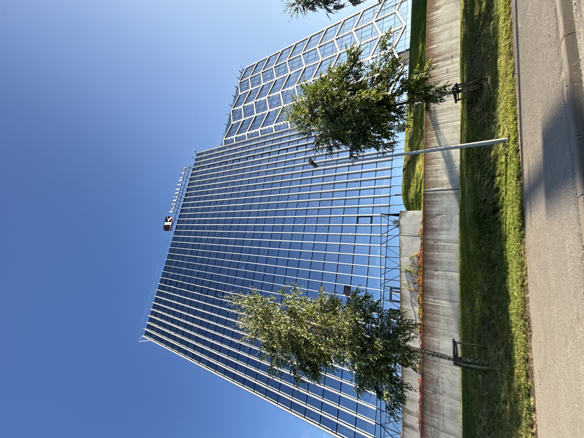 The image shows a modern glass-facade office building with the "Kaamos" logo visible at the top. The building is located in Tallinn, Estonia. In front of the building, there is a concrete wall and a grassy slope. Two trees are visible in the foreground, one on the left and another on the right. A street lamp is positioned to the right of the left tree. The sky is clear and blue.