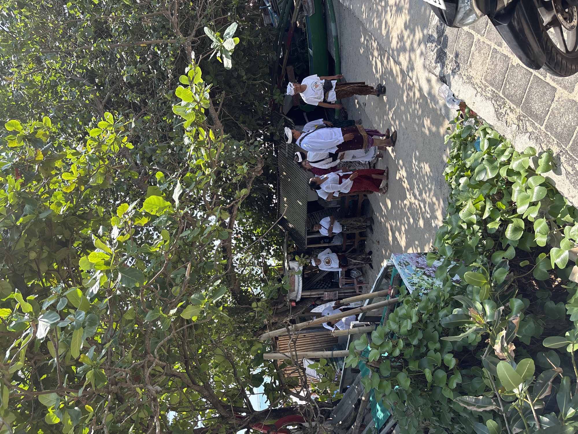 The scene is a street in Kuta, Indonesia. A group of people dressed in traditional Balinese attire are standing on a paved sidewalk next to a building. The clothing typically consists of a white top, head covering, and a sarong. Trees are visible on both sides of the street, with a motorcycle parked near the sidewalk's edge. The street appears to be in a shaded area. The light appears to be filtered through the trees.