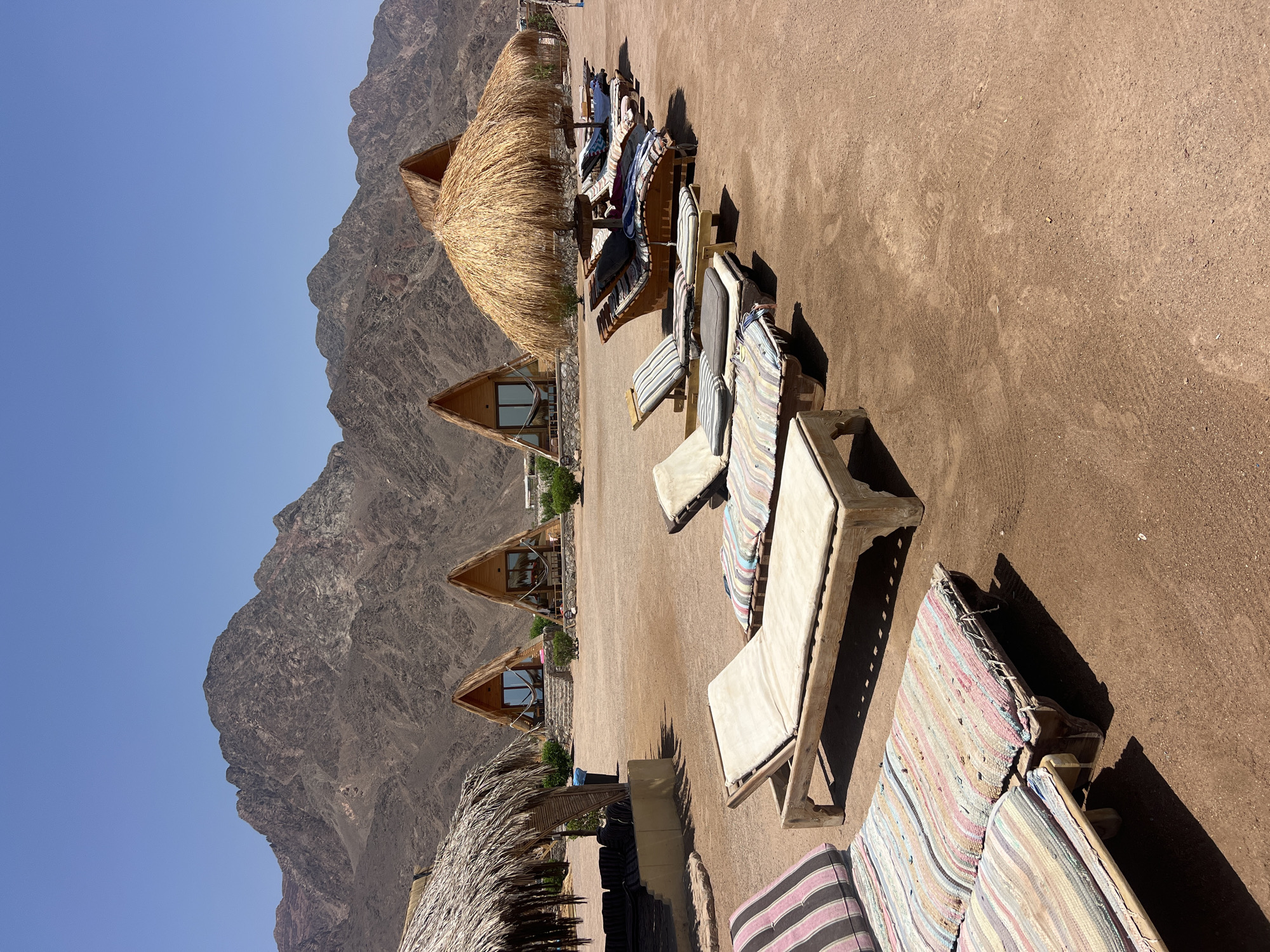 This is a wide, eye-level outdoor shot on a sunny day in Nuweiba, Egypt. The scene depicts a beach resort with triangular cabins with thatched roofs in the background. In the foreground, several wooden lounge chairs with cushions are arranged on the sandy ground. Some of the chairs are covered by a large thatched umbrella. A few sparse green bushes dot the landscape. In the distance, arid, rocky mountains rise against a clear blue sky. The ground is a light brown, sandy soil, with tire tracks visible. There are no people in the frame, and no action is taking place.