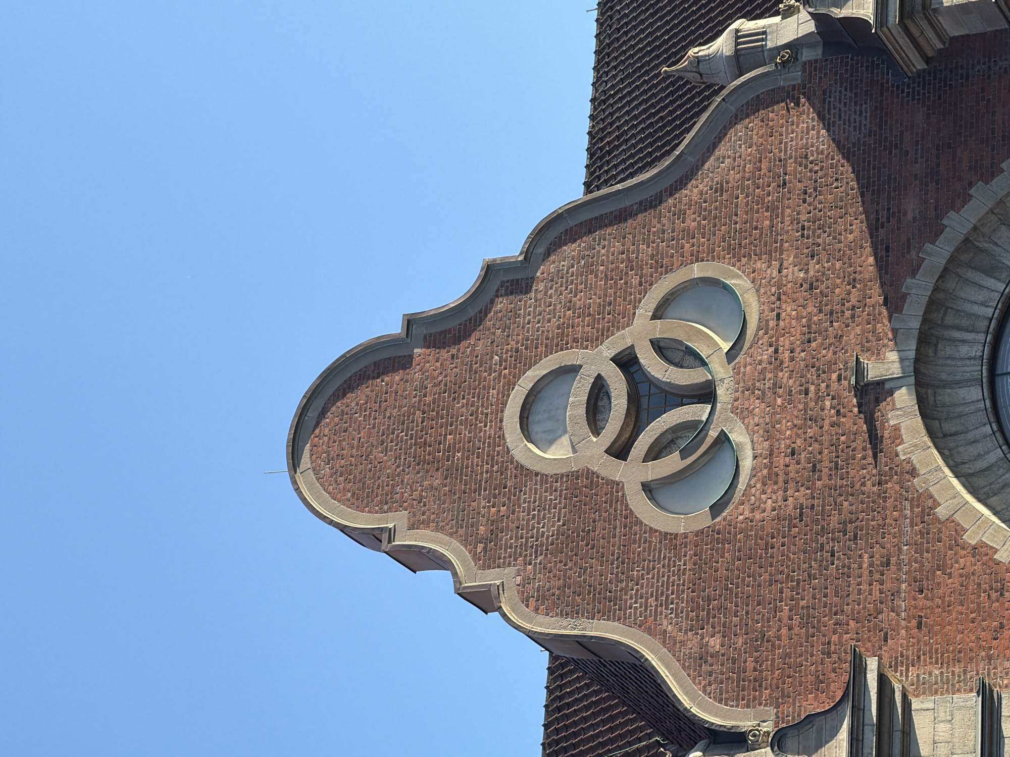The image shows the upper facade of a brick building, likely a historical structure, against a clear blue sky. The building features a decorative gable with a light-colored stone border. Centered on the gable is an ornate circular design composed of three interlocking rings, each containing a small window. The roof tiles are dark and have a visible texture. The lower portion of the image shows more of the building's facade, including arched stone structures. The overall impression is of architectural detail and historical character. This building is the former main entrance building of Malmö Museum.