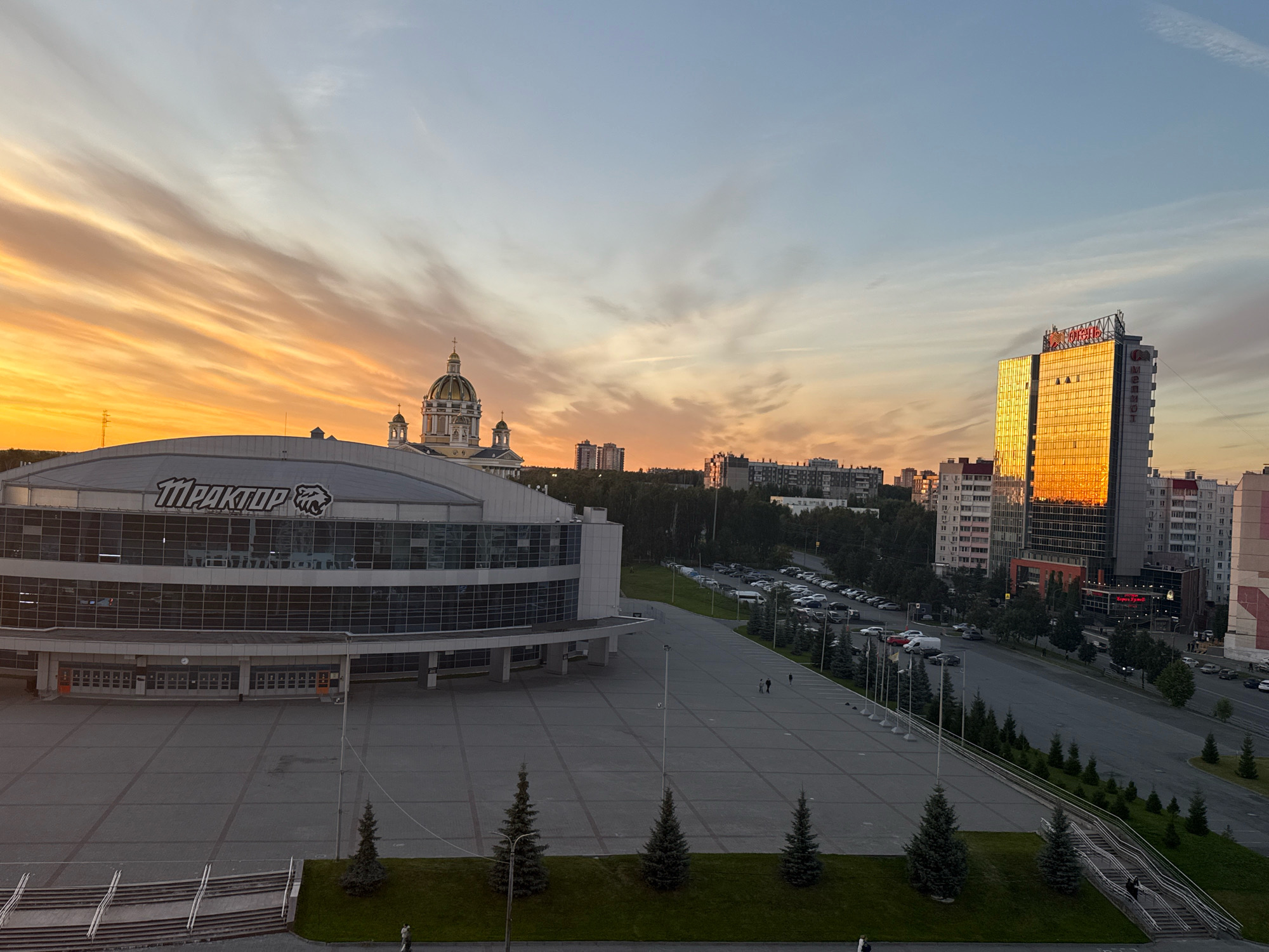 This is an eye-level, wide-angle photograph of the Traktor Ice Arena complex in Chelyabinsk, Russia, taken during a sunset. The sky is filled with wispy clouds, illuminated by the orange and yellow hues of the setting sun. In the foreground, a large, modern ice arena with a curved glass facade is visible, bearing the "TRAKTOR" logo. In front of the arena is a vast, paved plaza with several small evergreen trees planted in grassy areas along its perimeter. A long staircase with handrails leads down from the plaza to a lower level on the right side of the image.
In the mid-ground and background, a city skyline is visible. A prominent golden skyscraper reflects the sunset, and further to the left, a domed building, likely a church, stands out. A street lined with cars and trees runs alongside the arena and extends into the distance, where more residential buildings and some forest can be seen.
Two individuals are walking on the plaza near the arena entrances. On the street, several cars are parked and driving. On the staircase on the right, one person is descending. The overall scene suggests a tranquil evening in an urban environment.