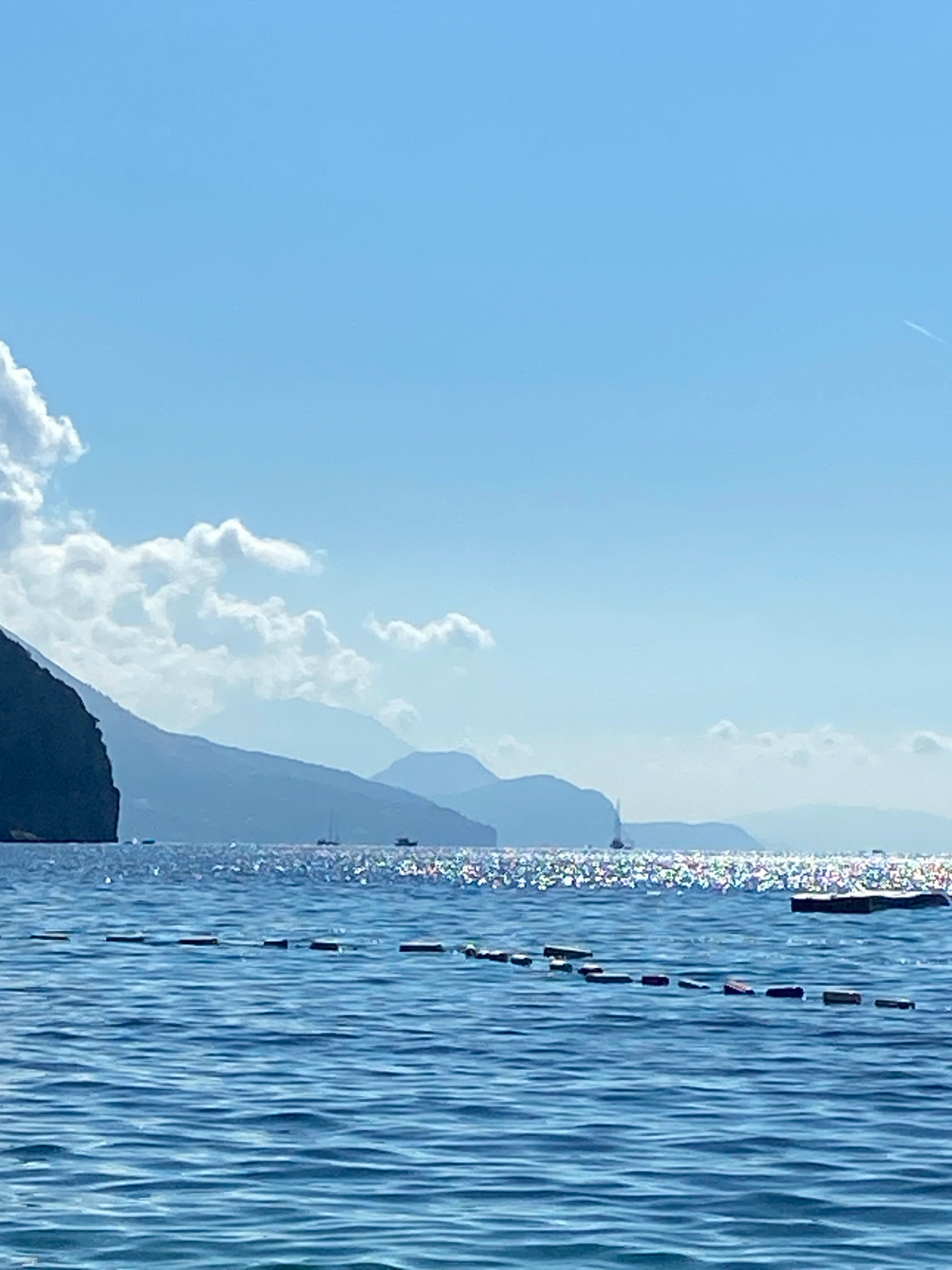 The image depicts a tranquil seascape with a clear blue sky and distant mountains. In the foreground, the ocean surface is rendered in shades of blue with visible ripples and sunlit patches. A line of buoys or markers stretches across the water from the bottom left towards the center. In the middle ground, several sailboats are visible on the sparkling water, and further back, the silhouettes of lush, mountainous islands rise against the horizon. The sky is a vibrant blue, with a few scattered white clouds near the left edge, and a faint contrail from an aircraft is visible in the upper right corner. There are no people or discernible structures in the scene, suggesting a natural coastal environment. The image does not provide any specific visual cues to pinpoint the exact location within Prijevor, Montenegro.