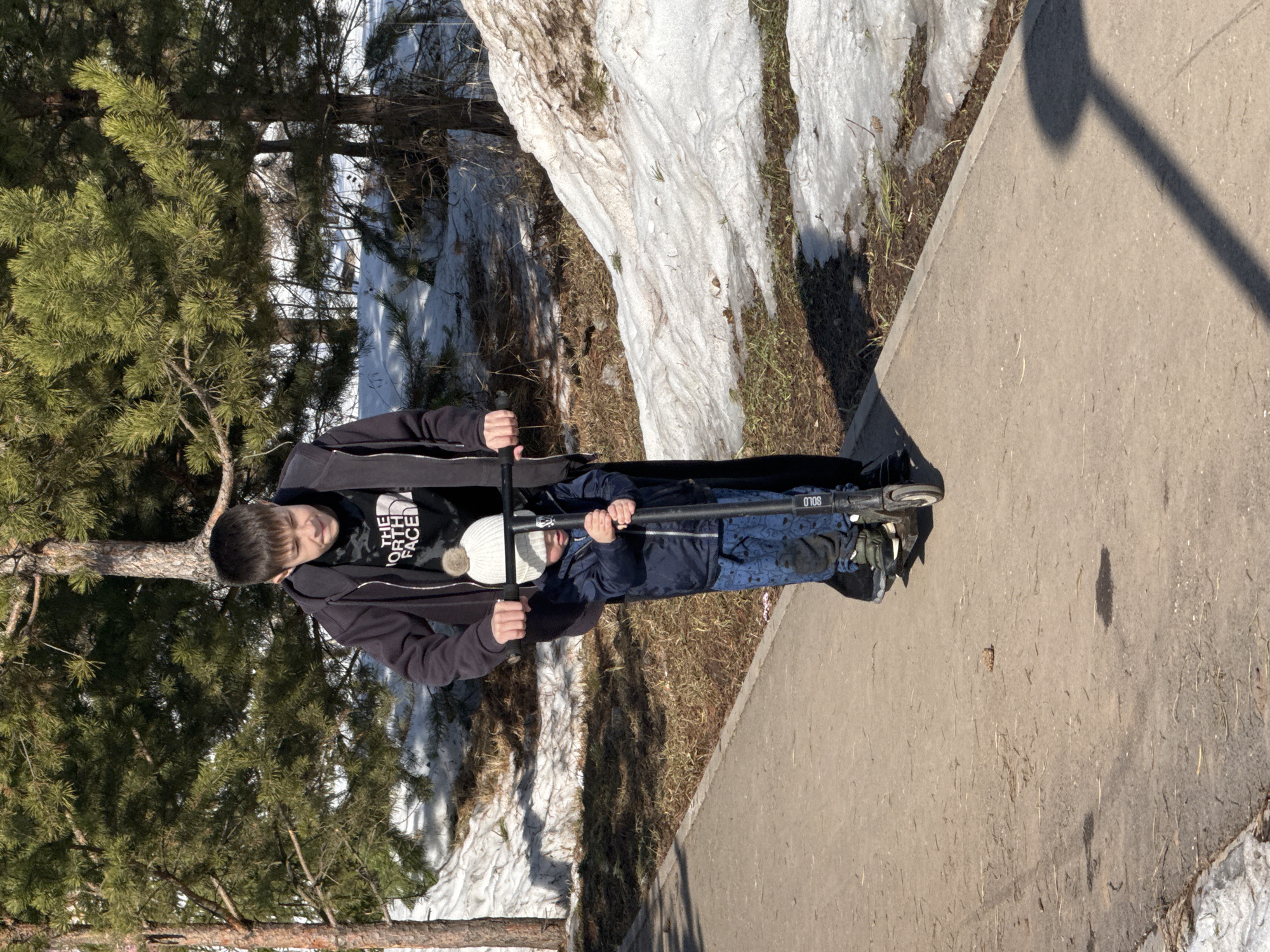 A teenager in a black jacket and North Face t-shirt stands on a concrete path holding a scooter with a small child in a blue jacket and white hat in front of him. The child is holding the scooter handle. To the left is a pine tree and snow-covered ground. To the right is a concrete path and snow-covered ground. The location is Zav'yalovo, Russia.