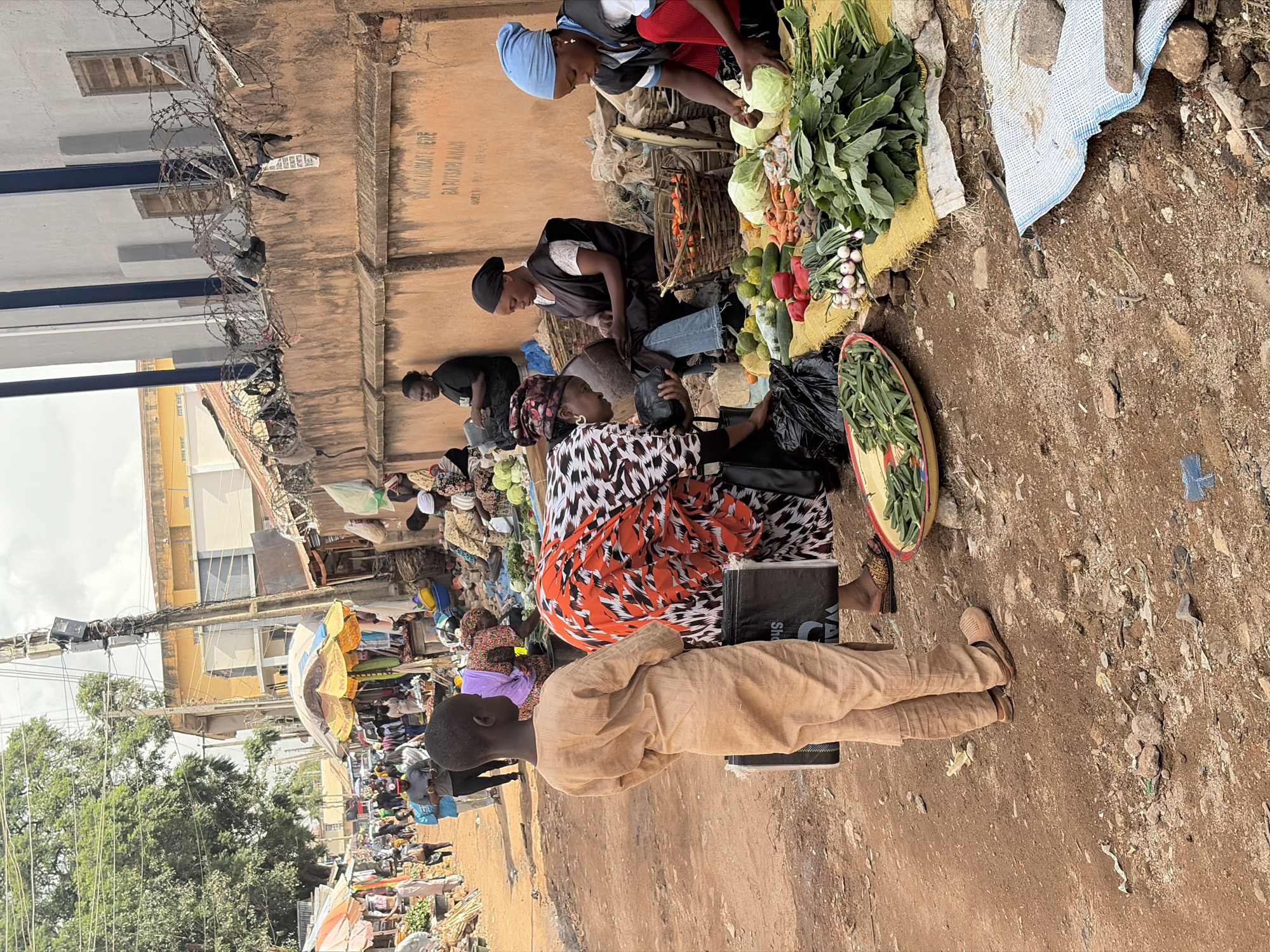 This image depicts a bustling street market scene in Jos, Nigeria, likely in an open-air area. The ground is a reddish-brown dirt, with scattered rocks and debris, indicating a somewhat unpaved environment. On the right side of the frame, several women are engaged in selling produce. One woman, wearing a blue headscarf, is arranging cabbages, while another, in a black headscarf, is tending to a basket of carrots and other vegetables. A third woman, in a vibrant orange and black patterned top, is interacting with a customer who is partially visible, holding a black bag. In the foreground, a young boy in a light-colored tunic stands looking away from the camera, holding a similar black bag.
The market appears to be set up along a wall, possibly part of a building, which is made of weathered concrete or brick. Barbed wire is strung along the top of this wall, suggesting a security measure or boundary. Further down the street, more stalls are visible under colorful umbrellas, with various people browsing and interacting. Utility poles with numerous wires are prominent in the background, typical of urban infrastructure. The overall atmosphere suggests a lively and active marketplace where daily commerce is taking place.