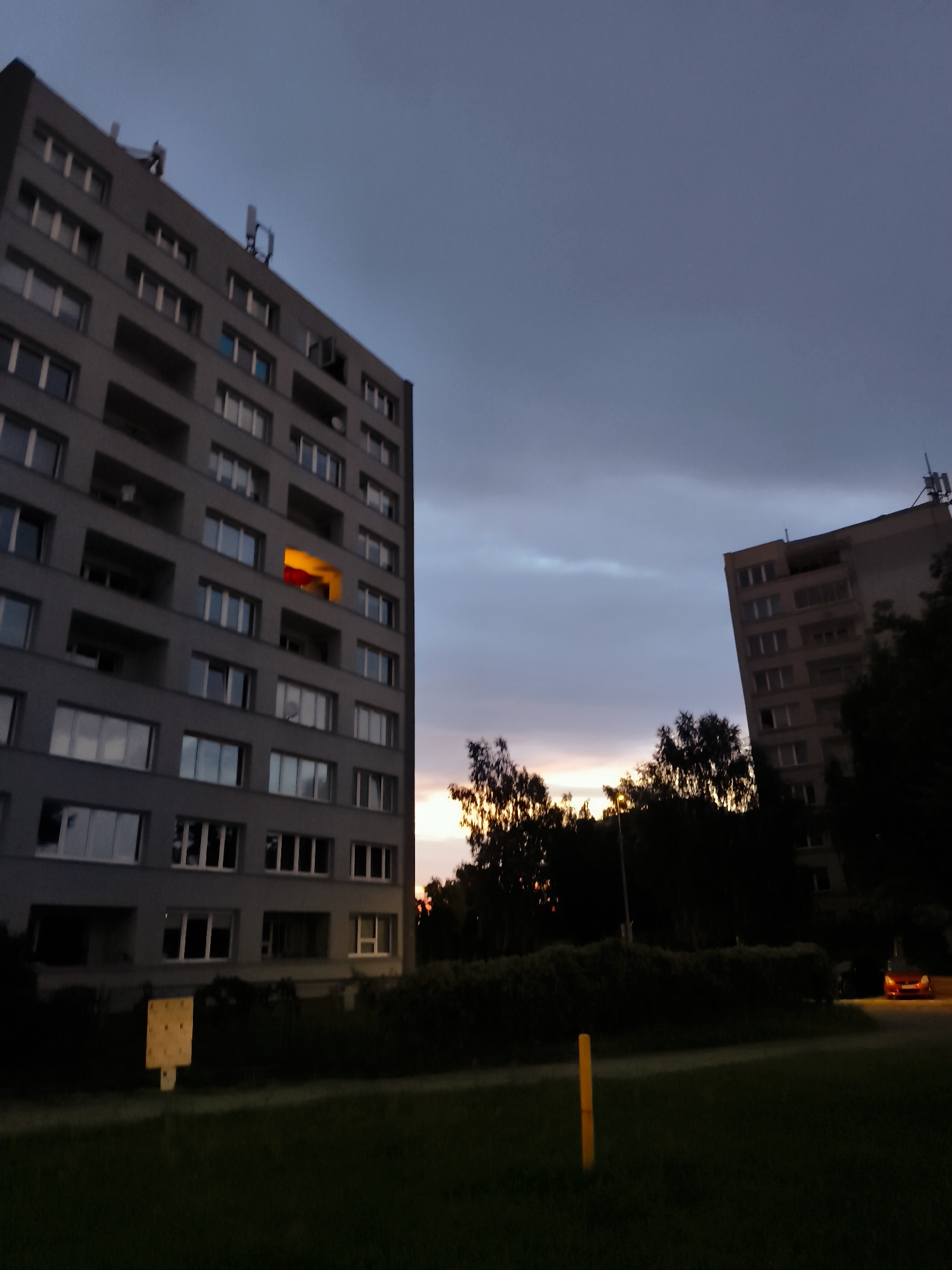 The image depicts a twilight scene in Gdańsk, Poland, featuring two tall, gray residential buildings. The building on the left is closer and larger, with numerous windows and balconies. One window on this building is illuminated with a warm orange light, suggesting occupancy. Antennas are visible on the roof of the building. The building on the right is further away and appears slightly lighter in color. A dense line of dark trees separates the buildings from a grassy foreground. A red car is parked in the background near the building on the right, its headlights on. The sky is a muted, cloudy gray with hints of orange and pink from the setting sun visible through the trees. A yellow marker post stands in the grassy area in the foreground. The overall atmosphere is serene and suburban.