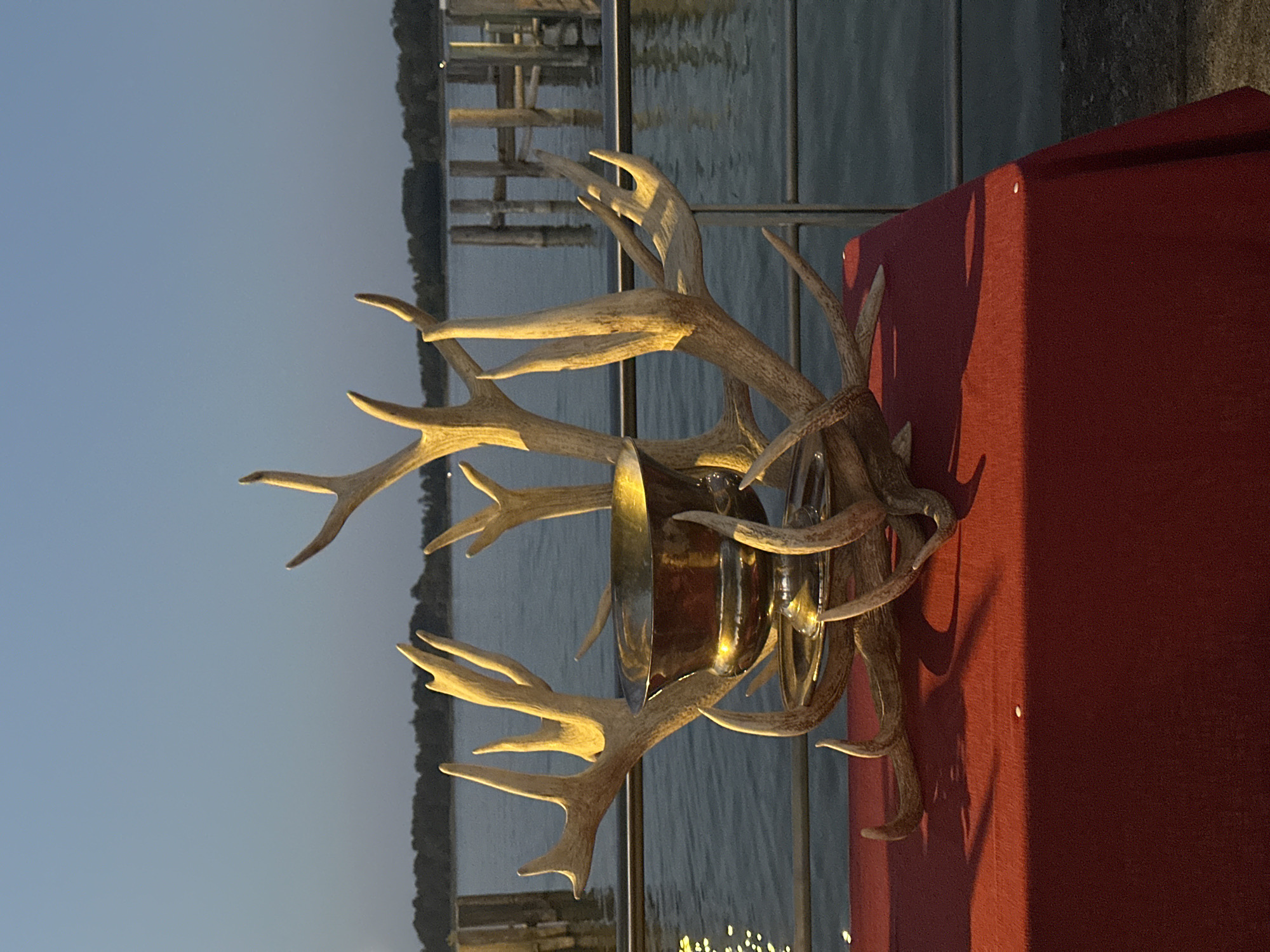 The image depicts a trophy made of antlers and a metal bowl, displayed on a red tablecloth. The trophy is placed on a table with a railing and a body of water in the background. Several wooden pilings are visible in the water, suggesting a waterfront location such as a pier or marina. The sky is clear and dusky, indicating either sunrise or sunset. The overall scene is an outdoor setting near a body of water, possibly in Prien am Chiemsee, Germany, given the context provided. There are no people visible in the image.
