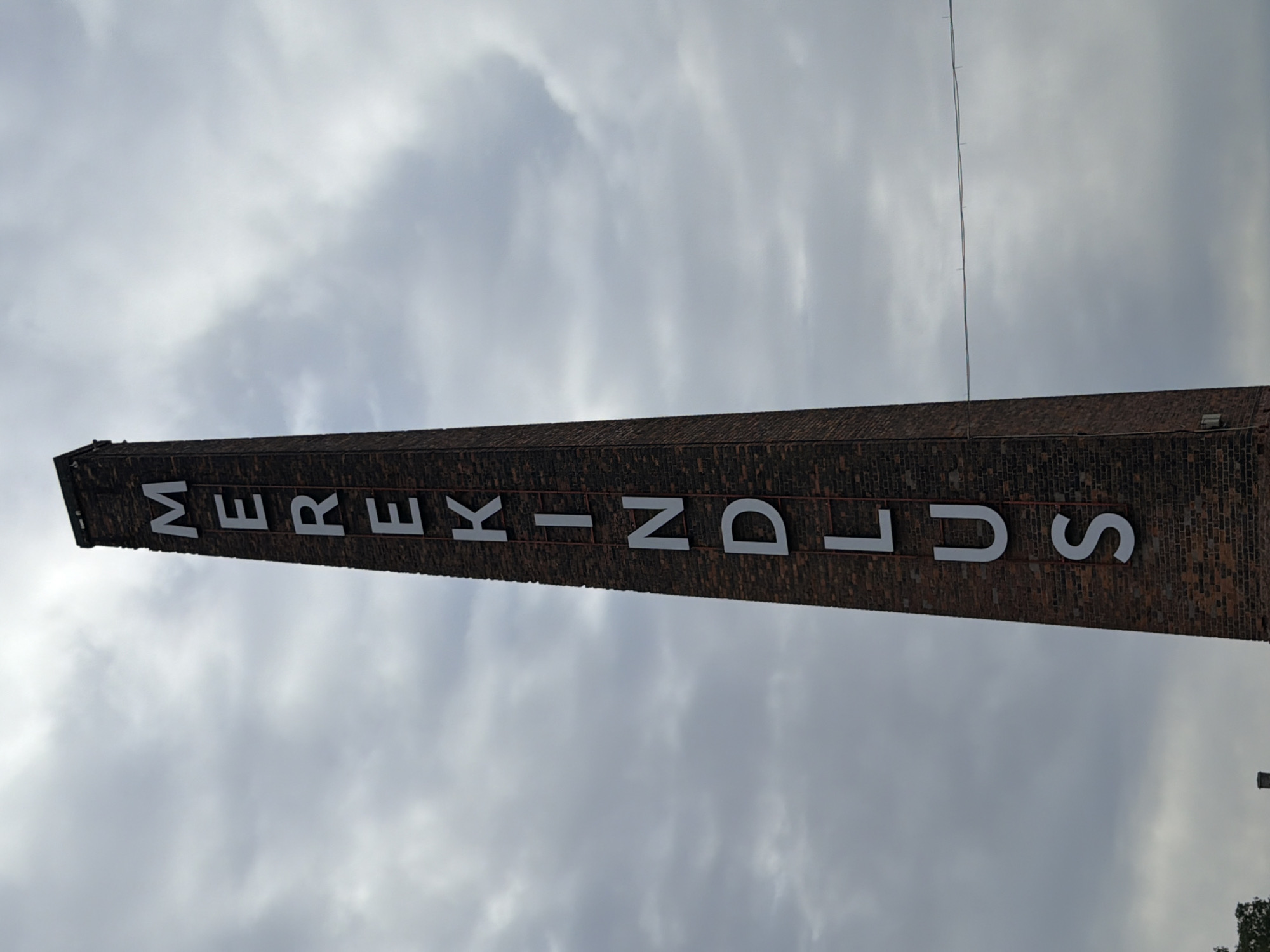This image shows a tall, brick chimney with the word "MEREKINDLUS" written vertically in large, white letters. The chimney is located in Tallinn, Estonia. The sky is cloudy and overcast. There are no people visible in the image. The chimney appears to be an industrial structure.