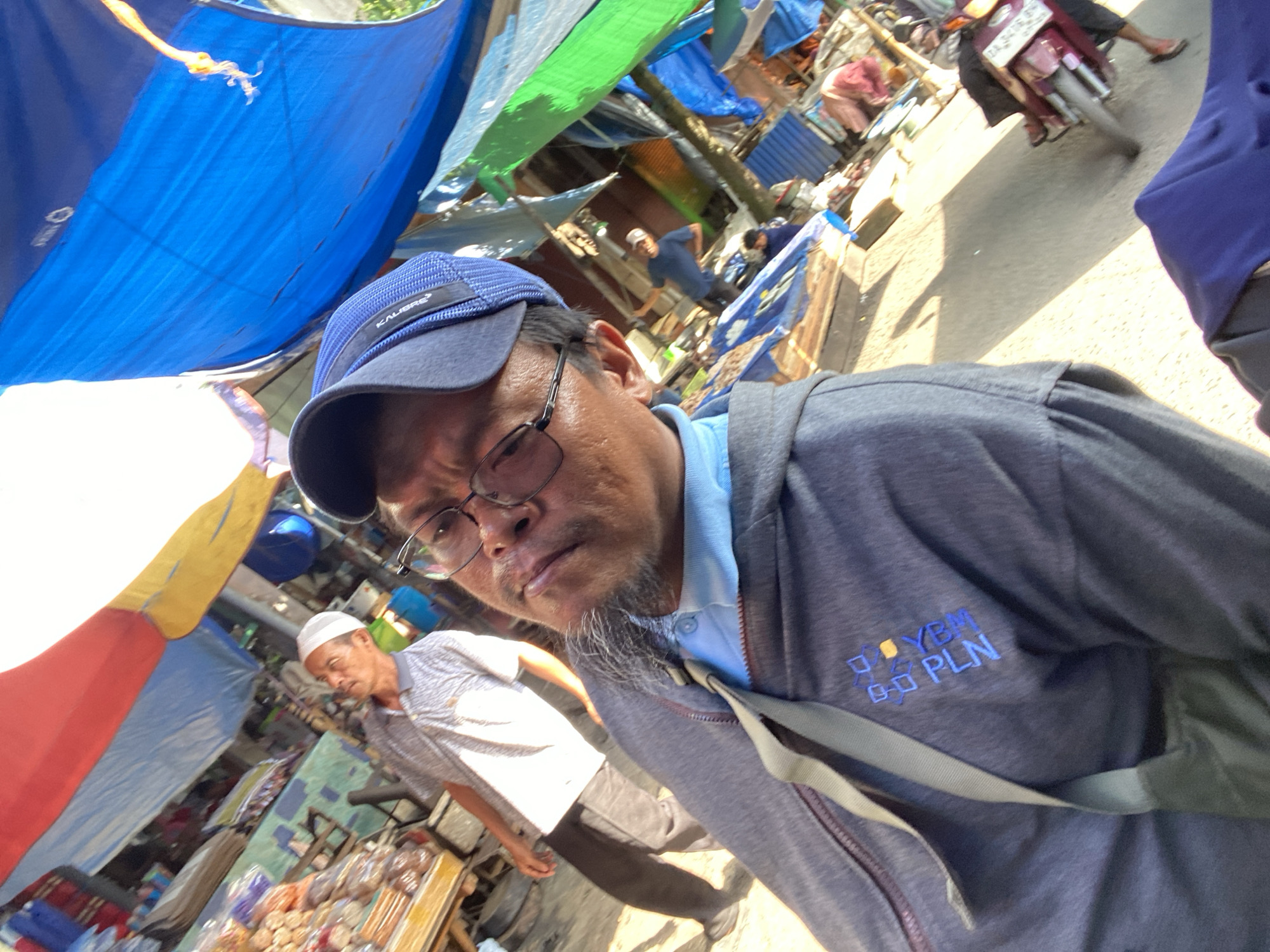 A middle-aged man wearing glasses and a blue baseball cap takes a selfie at an outdoor market. He is wearing a grey hooded sweatshirt over a light blue collared shirt, and a grey strap is visible across his chest. The background shows a bustling market scene with stalls covered by colorful tarps, including red, yellow, and blue. Another man in a white cap and grey shirt is visible in the background, as well as a motorcycle and other marketgoers. The overall lighting suggests it is daytime. The image was taken in Desa Tunggul Irang, Indonesia.
