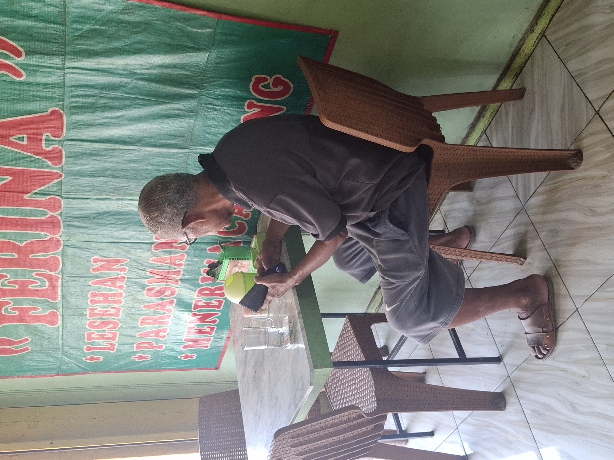 An elderly man with gray hair and glasses is sitting at a marble-topped table in a restaurant, looking at his phone. He is wearing a dark gray polo shirt and gray shorts, with brown leather sandals on his feet. The table has a glass of water, a pack of cigarettes, and a mobile phone on it. To the left of the man, a green basket holds utensils. In the background, a large green banner with red lettering reads "FERINA" and below it, "LESEHAN PARASMAN MENERIMA TAMU" (which translates to "LESEHAN PARASMAN ACCEPTS GUESTS" and likely refers to a type of seating and a restaurant or establishment). The man is seated in a brown plastic chair, and another similar chair is visible to his left. The floor is tiled with a light-colored pattern. The walls appear to be painted a light green color. The scene is set in the city of Cibadak, Indonesia.