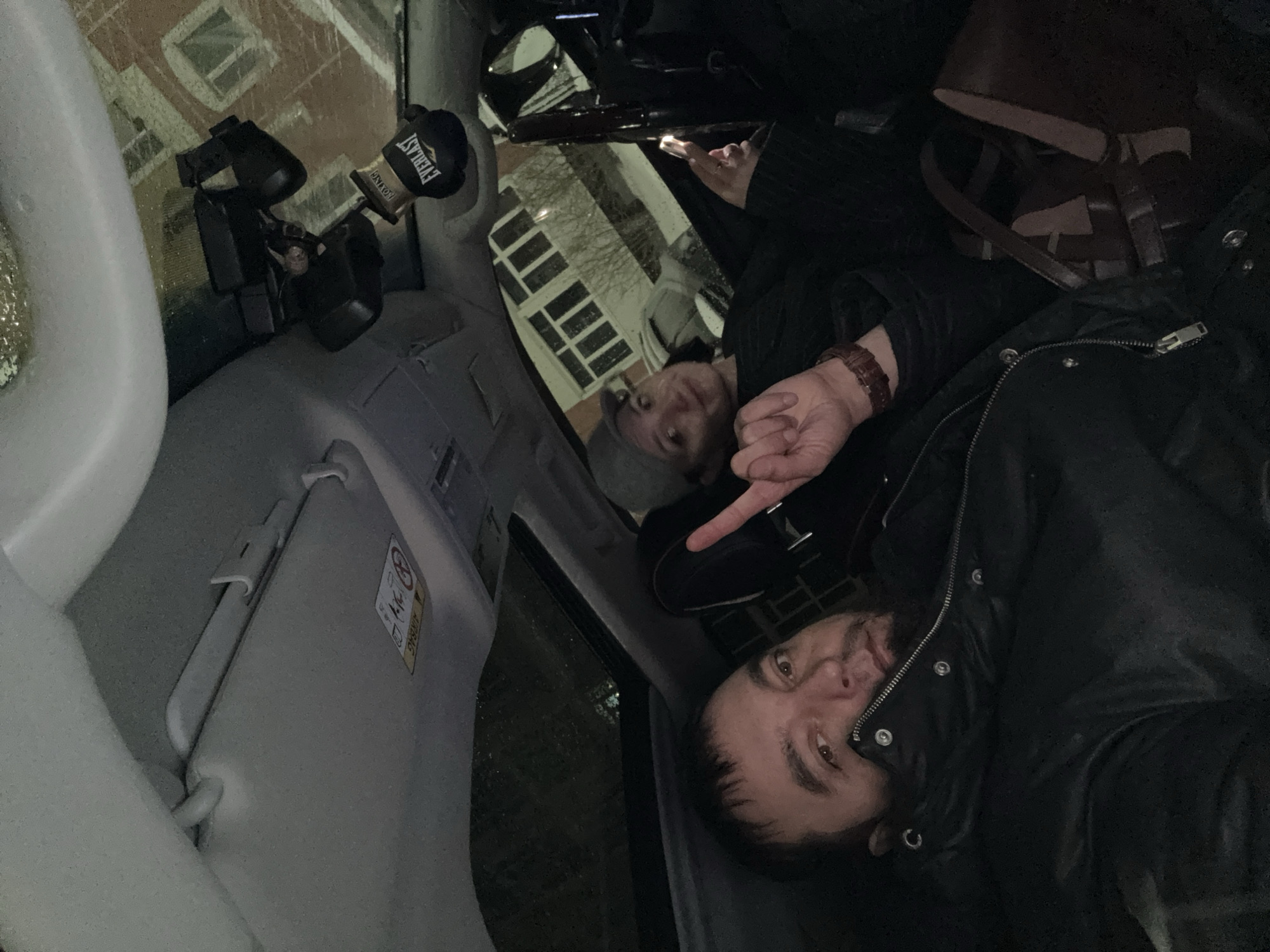 A low-angle shot inside what appears to be a vehicle, revealing parts of the interior, a partial view of two people, and a glimpse of the outside environment.
Interior: The vehicle's interior includes a light-colored visor with an airbag warning label, a black car phone holder with a pair of black binoculars, and what appears to be a small Everlast boxing glove.
People: Two people are partially visible. A man with short dark hair is closer to the foreground wearing a black jacket with the zipper partially visible. The second person, possibly a woman, is located behind him wearing a gray hat and black jacket, partially obscured. One person is pointing towards the other.
Environment: A glimpse of a building with multiple windows and a street outside suggests an urban environment. The building facade and general atmosphere appear consistent with an urban setting.
Actions/Interactions: The two individuals seem to be interacting within the confined space of the vehicle.
The presence of the binoculars suggests possible observational activities. The picture appears to be taken inside a vehicle in Nizhnevartovsk, Russia.