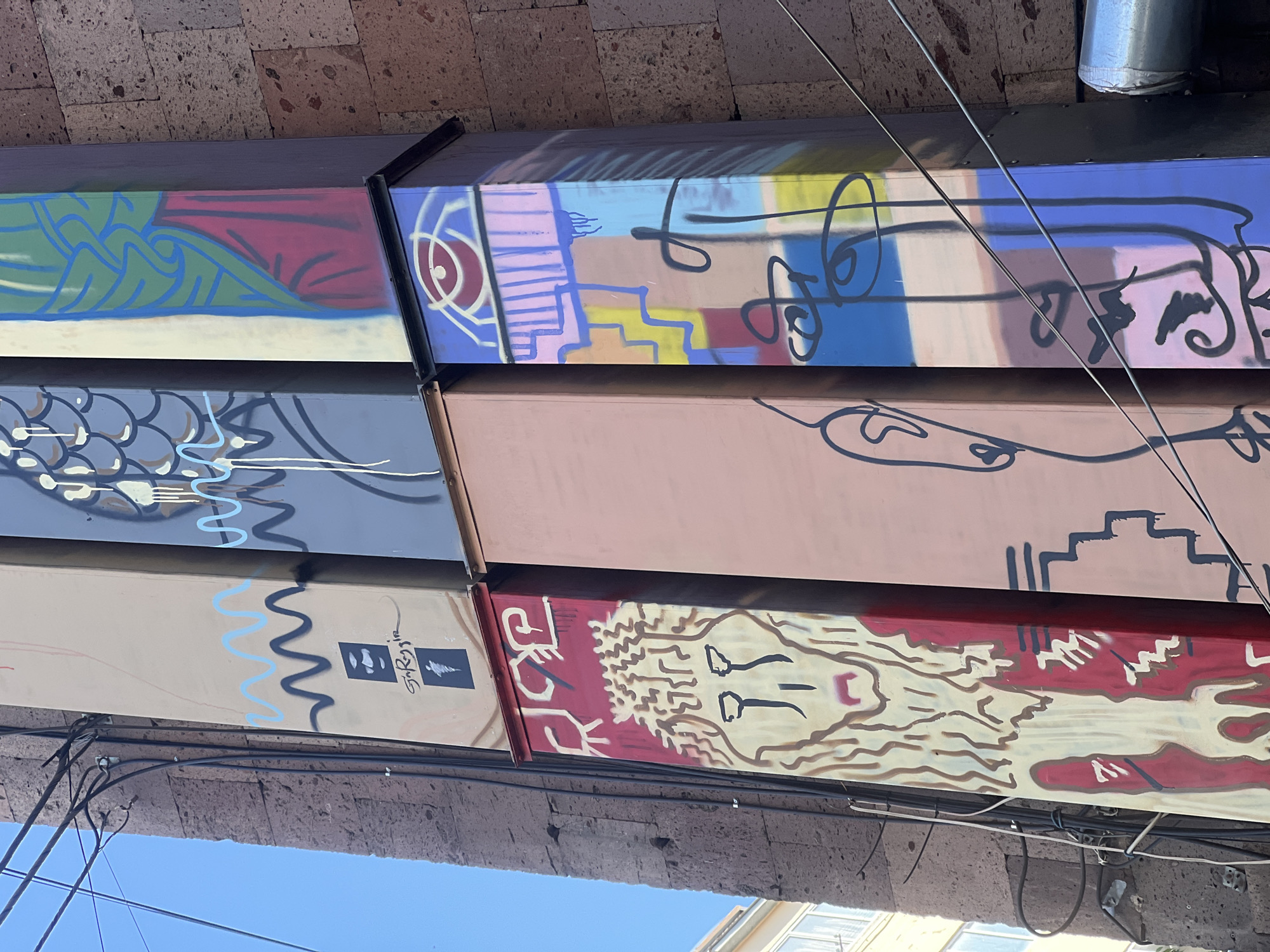Captured from a low angle in Yerevan, Armenia, the image features a row of painted wooden beams, likely part of a building's structure or an art installation. Each beam showcases a different abstract design or portrait, employing a range of colors and styles. A mix of urban infrastructure, including brickwork, wires, and parts of other buildings, fills the peripheral spaces of the frame. The sky is visible in the lower portion of the image.