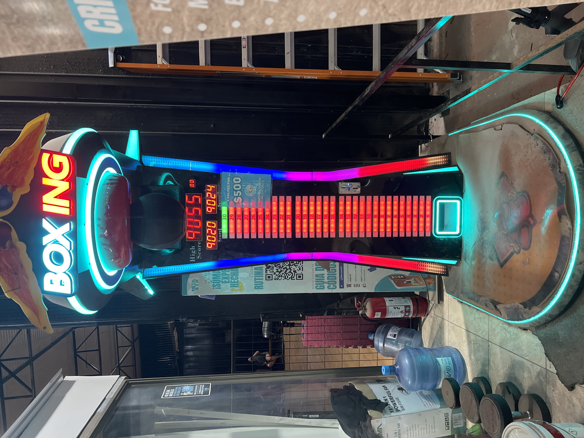 The image captures a boxing arcade game, likely located in Arica, Chile. The game features a neon-lit design with a prominent "BOXING" sign. The game's display shows high scores: 9055, 9020, and 9024. Beneath the display are instructional signs in Spanish, including one mentioning a prize of $500 and another with a QR code labeled "RUTINA". The surrounding environment includes a partially visible storefront window, metal fencing, water jugs, a fire extinguisher, weights, and boxes labeled "POWERFUL FAN". A person is faintly visible behind the metal fencing. Ladders are visible near the top of the frame. The ground appears to be tiled.