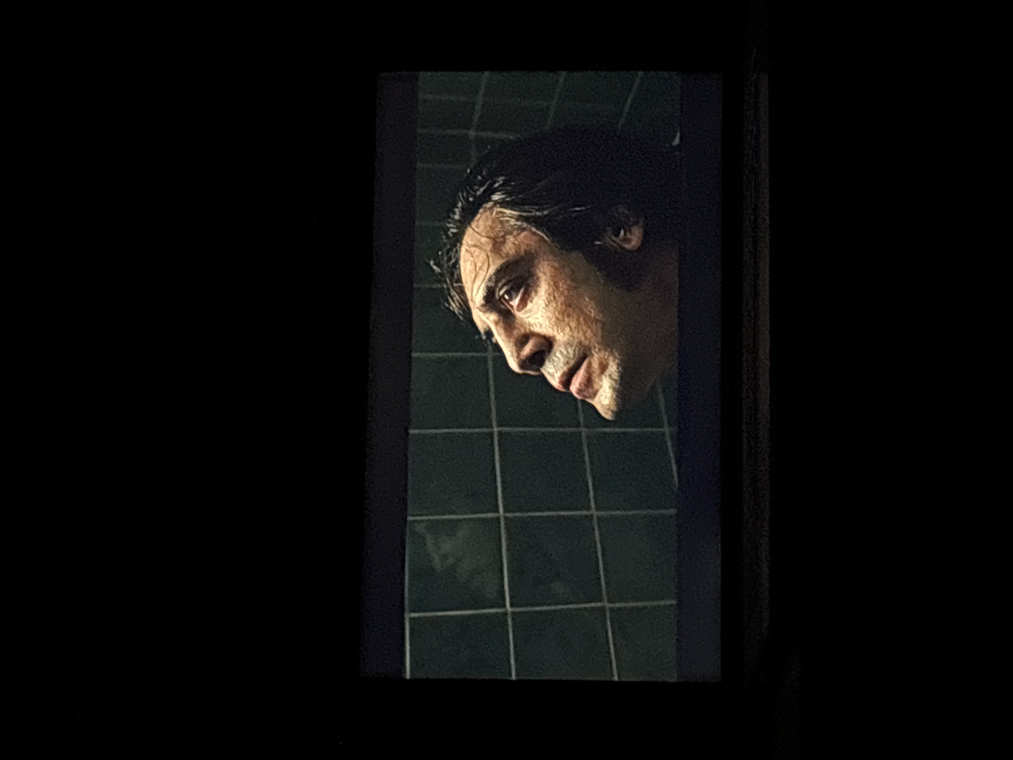 The image displays a still frame from a film or television program projected onto a rectangular screen, viewed in a dark environment. The central subject within the screen is a man positioned in profile, looking upwards and slightly to his left. He has dark, wet hair that is matted against his forehead and temples. His face shows detailed texture, including visible pores and some stubble along the jawline and chin. His expression appears contemplative or pained, with his mouth slightly agape. The background of the projected scene consists of dark teal or green square tiles, suggesting an indoor setting like a bathroom or shower. The screen itself is bordered by absolute darkness, indicating the photograph was taken in a dim room. There are no identifiable elements or landmarks within this image that indicate a location in the city of Budva, Montenegro.