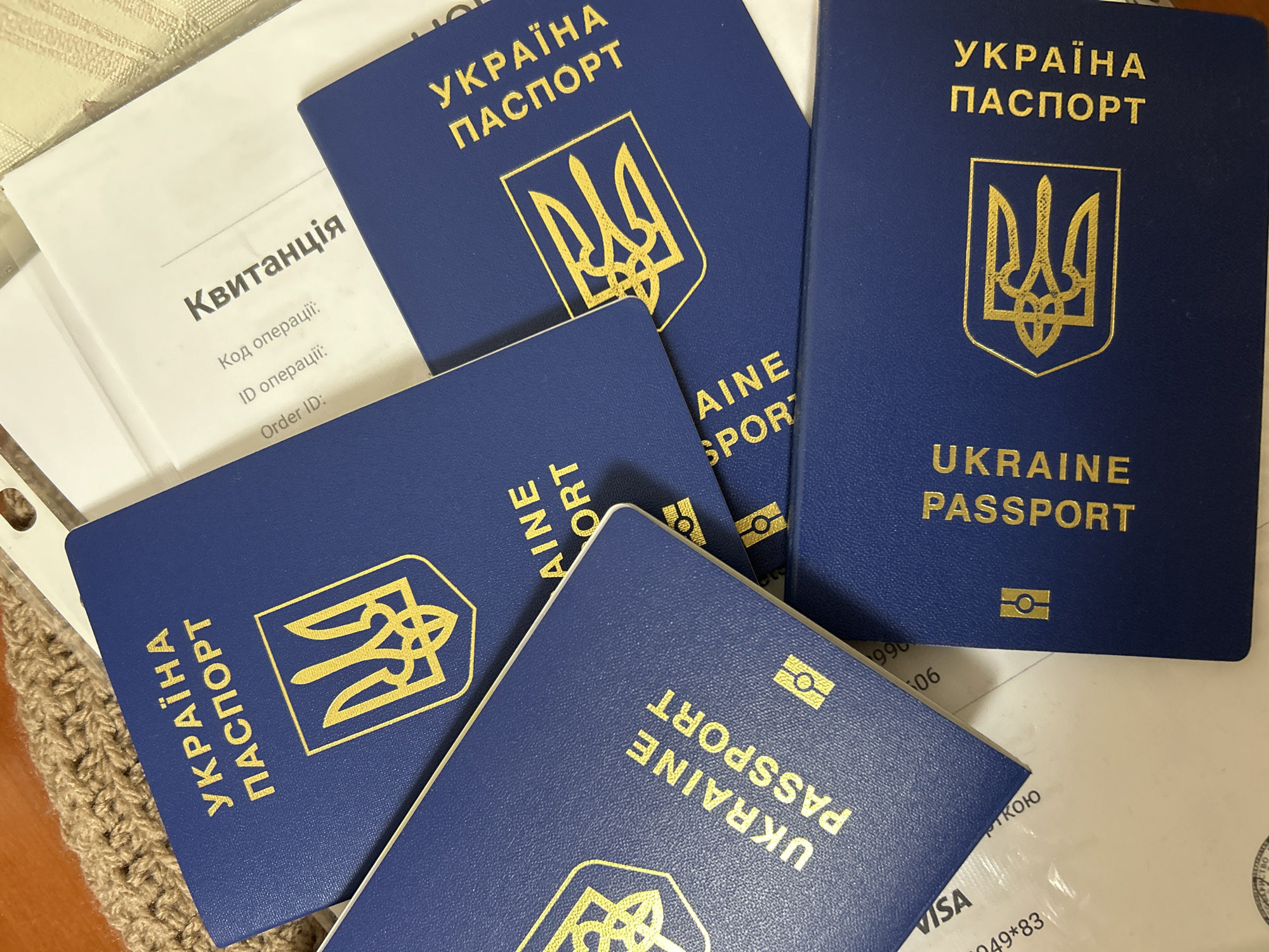 The image displays several Ukrainian passports and a document with Ukrainian text, including the word "Квитанція" (Receipt). The passports are dark blue with gold lettering and the Ukrainian coat of arms. They are arranged in a stack and overlapping. One passport shows "UKRAINE PASSPORT" in English. The document appears to be a printed form with lines for "Код операції" (Operation code) and "ID операції" (ID operation). The overall scene suggests a setting where official documents are being handled, possibly for processing or storage. The environment includes a textured surface, possibly a knitted item, and a white flat surface. There are no people visible in the image, and no specific actions or interactions are depicted. The location in Ivanivka, Ukraine is not visually evident from the provided image.