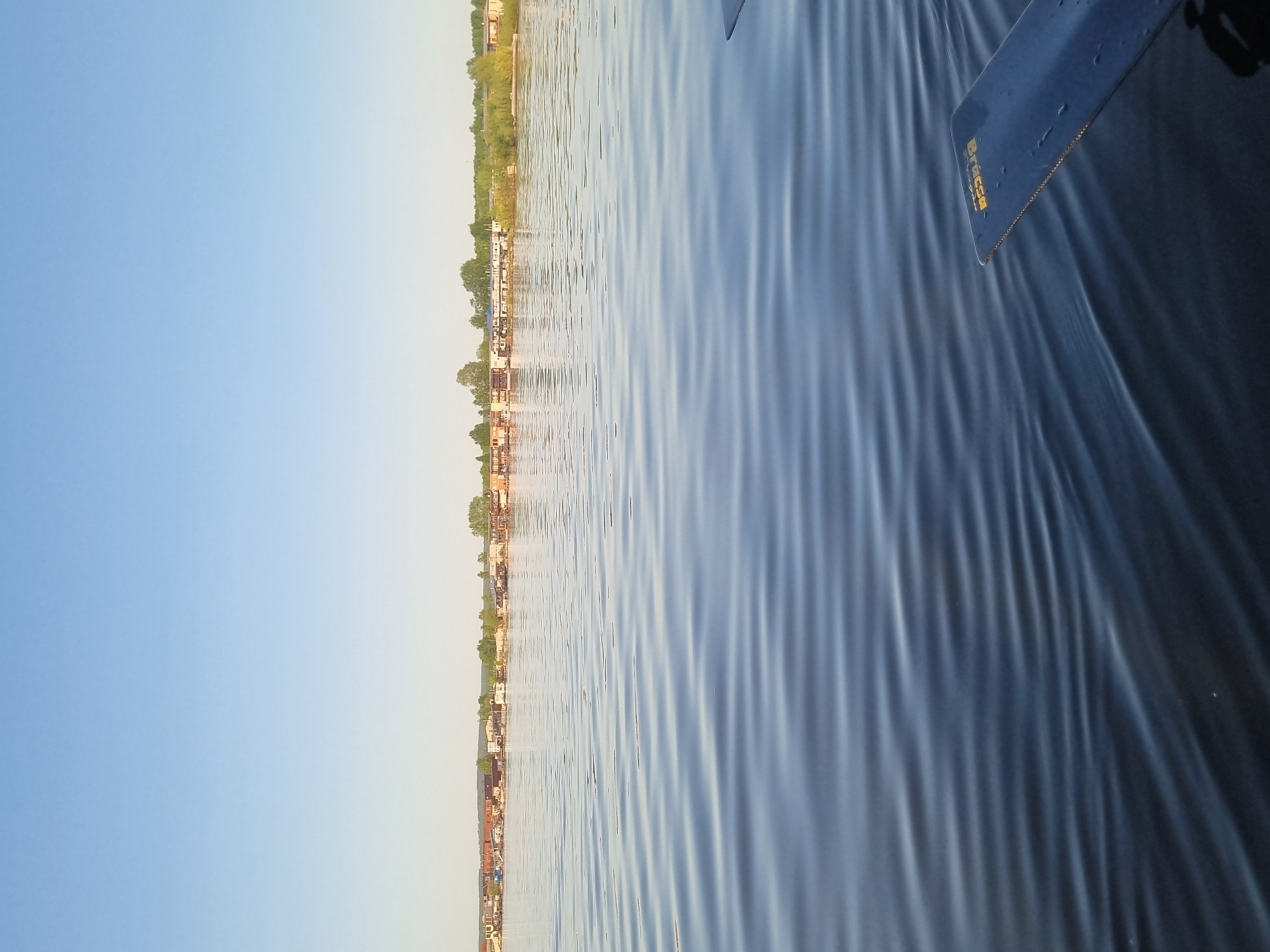 The image shows a body of water, likely the Kama River, with the city of Chaikovsky, Russia, visible in the background. The sky is clear and blue. In the foreground, part of a watercraft, possibly a boat or paddleboard, is visible. The water is rippled. The shoreline features vegetation and buildings.