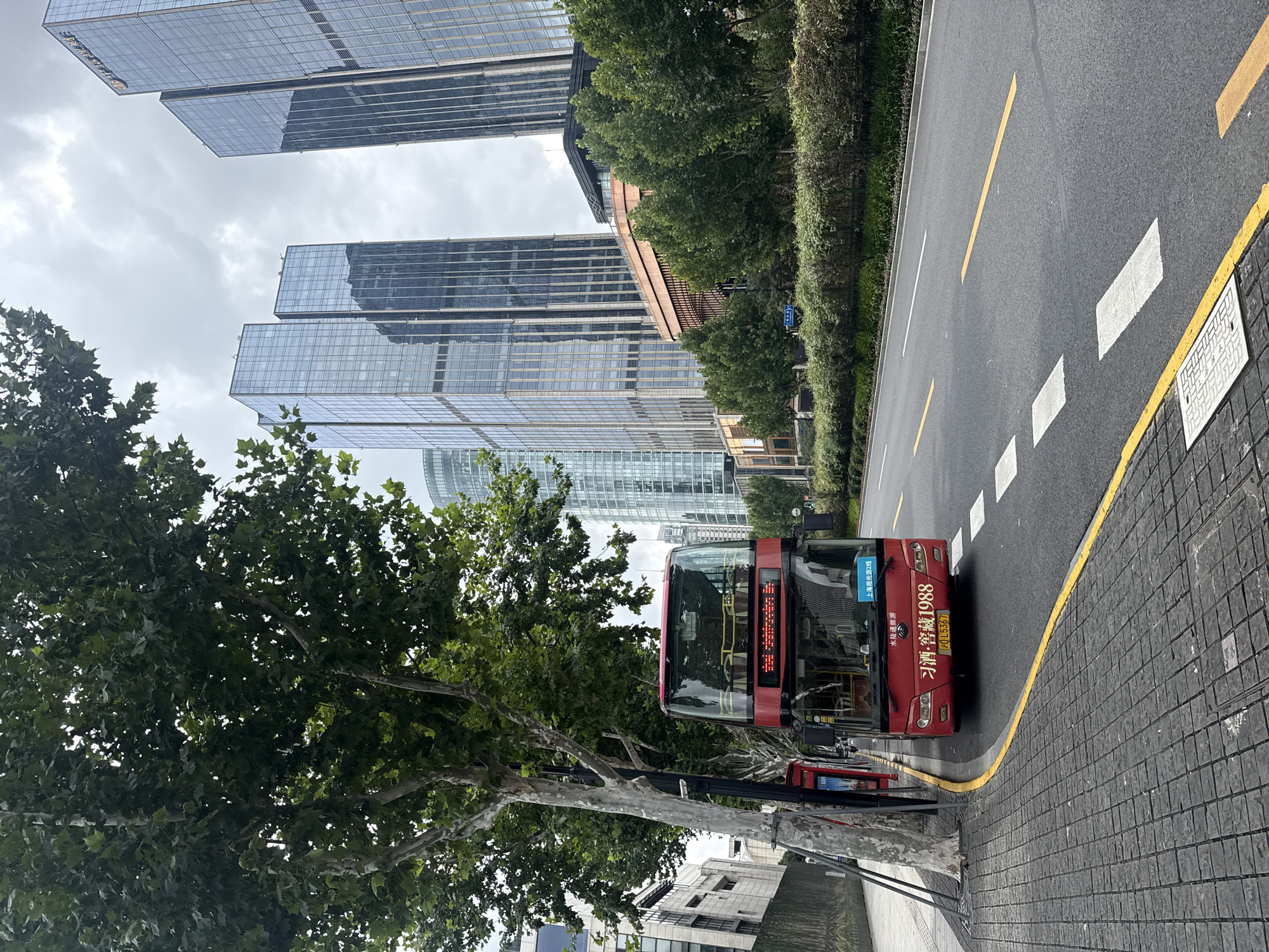 In Shanghai, China, a bright red double-decker bus is parked on the side of a street, its front facing the viewer. The bus has a digital display on its front showing "Shanghai Sightseeing Line 2". A license plate with the characters "鲁L5367" is visible. The bus is parked on a paved sidewalk next to a road with yellow and white lane markings. Tall, modern glass skyscrapers rise in the background, reflecting the overcast sky. Trees with lush green foliage line the street, casting shadows on the sidewalk. A few red telephone booths are also visible along the sidewalk. The scene depicts a typical urban environment in Shanghai with a blend of historical and modern architecture.