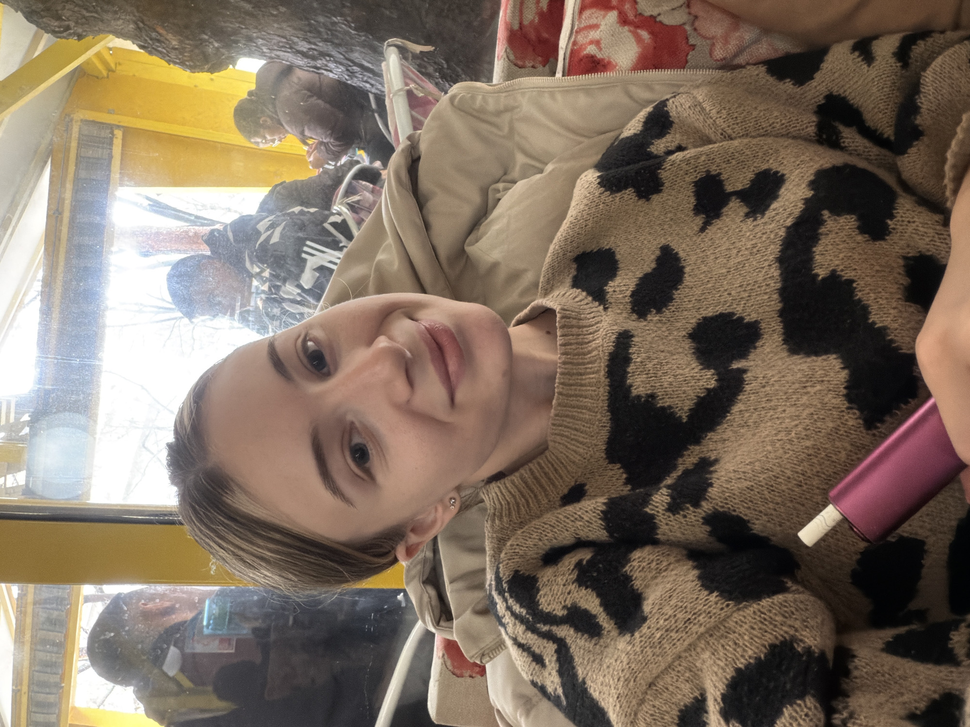 A young woman is in a glass-enclosed structure, potentially a gondola or observation pod, possibly in Plovdiv, Bulgaria, as it appears to be the city in the reflection. She wears a tan-colored sweater with large, dark brown or black animal print markings, and a light tan jacket. In her hand, she holds a small, cylindrical object that appears to be a heated tobacco product device with a white filter. Reflections in the glass show other people and trees. The structure has yellow-painted framing.
