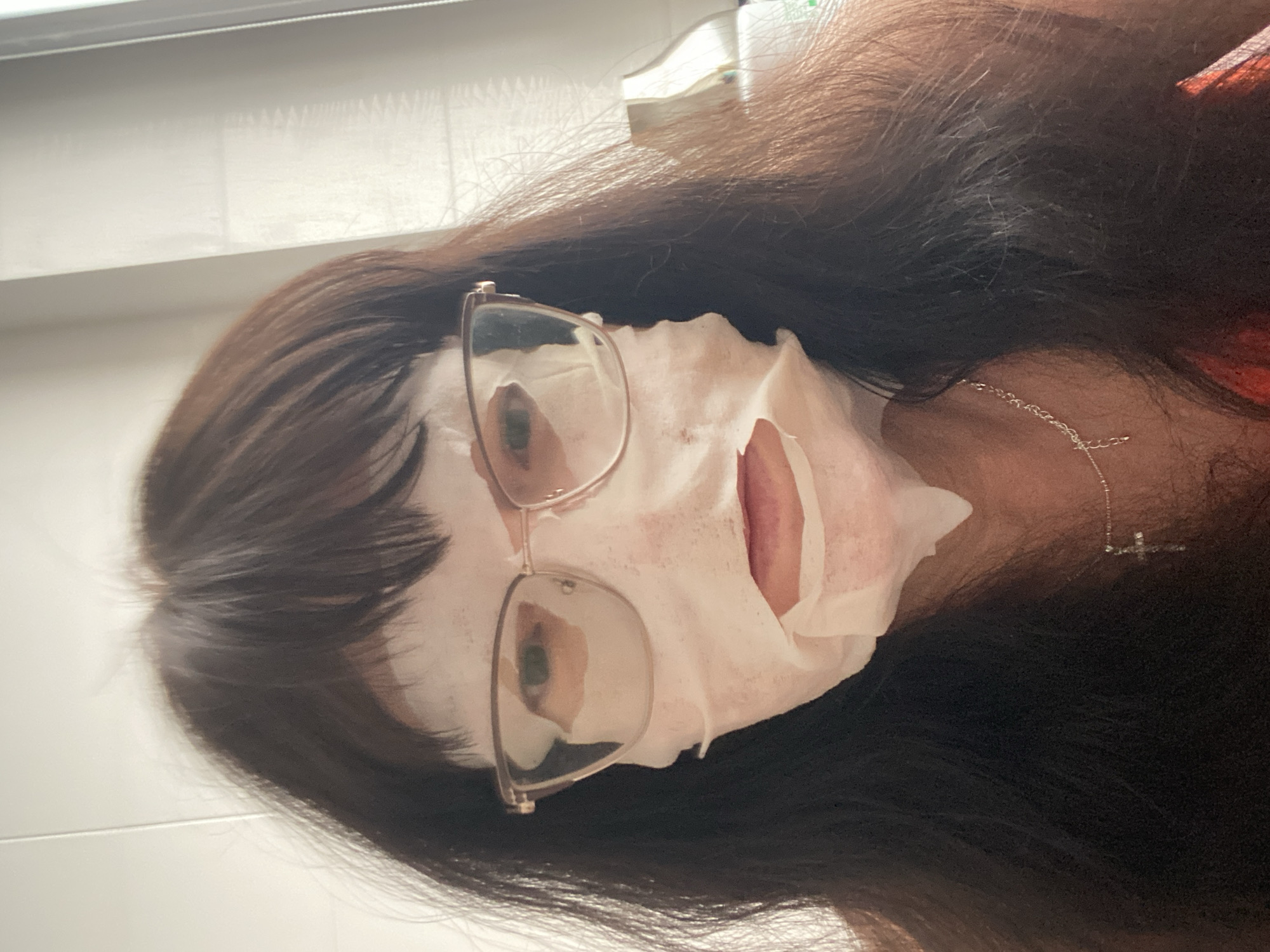 A person is wearing a white facial mask and glasses, with their dark hair framing their face. A silver necklace with a cross pendant is visible around their neck. The background appears to be indoors, with a white wall and a curtained window. The location is identified as Novi Petrivtsi, Ukraine.