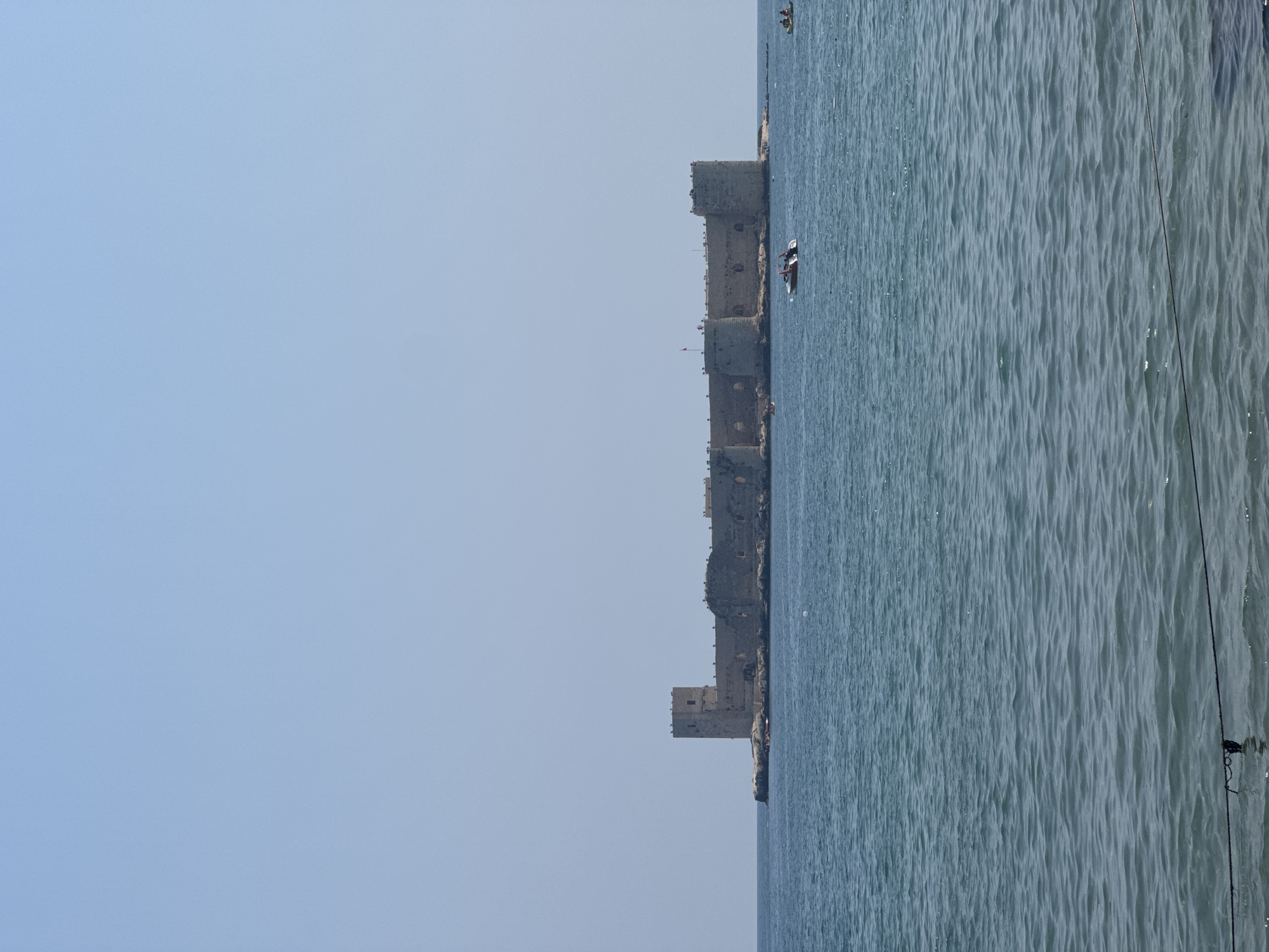 The image shows the Maiden's Castle (Kızkalesi) located off the coast of Erdemli, Mersin Province, Turkey. The castle is a historical stone fortress situated on a small island in the Mediterranean Sea. The sky is clear and blue, with a few very light clouds. The sea is a deep blue and appears calm with gentle ripples. A few boats are visible on the water, including a small motorboat with people on board near the castle, and what appear to be kayaks or similar small watercraft further in the distance. The castle itself has several towers and thick stone walls, with evidence of some reconstruction or preservation. The overall scene is one of a historical landmark situated in a coastal environment on a clear day.