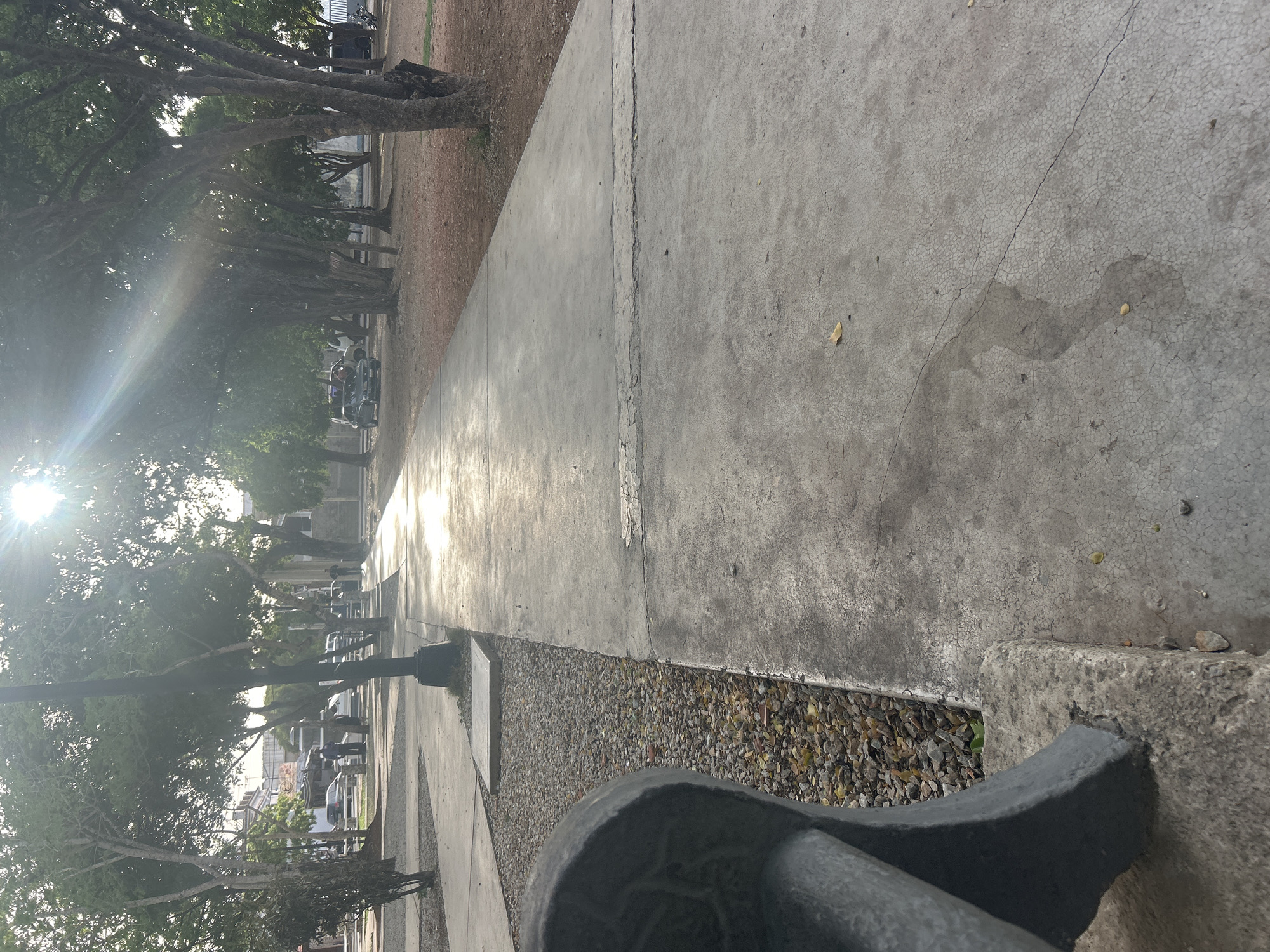 This is a ground-level, vertical-format photograph of a paved walkway in a park in Santa Ana, Venezuela. The walkway, made of concrete, is illuminated by bright sunlight filtering through the canopy of trees that line its sides. The sun's rays create a shimmering effect on the wet or glossy surface of the path. To the left of the walkway, a section of a dark-colored park bench is visible in the foreground, partially obscuring a gravel-covered area with scattered leaves. Beyond the walkway, on the right, is a dirt or mulch-covered area with trees. In the background, several vehicles are parked, and some individuals are present, walking or standing near the parked cars. The overall scene depicts a public park setting with natural elements and signs of human activity.