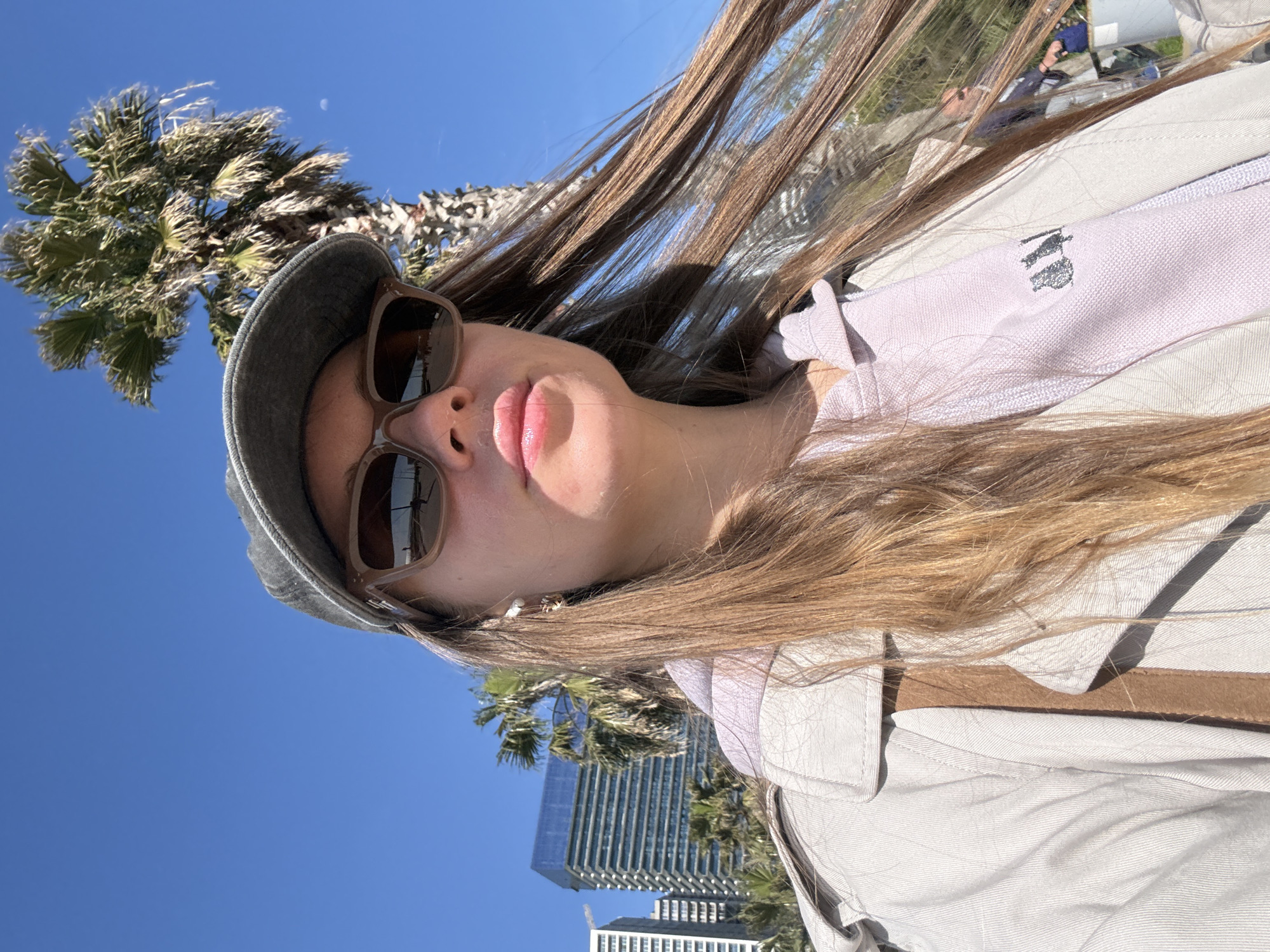 A woman wears sunglasses and a cap, posing in front of palm trees and buildings under a blue sky. The photo appears to be taken looking up at her. A tall building is in the background. Based on the architecture and the presence of palm trees, the image may be taken near the Batumi Boulevard in Batumi, Georgia, where such scenery is common.