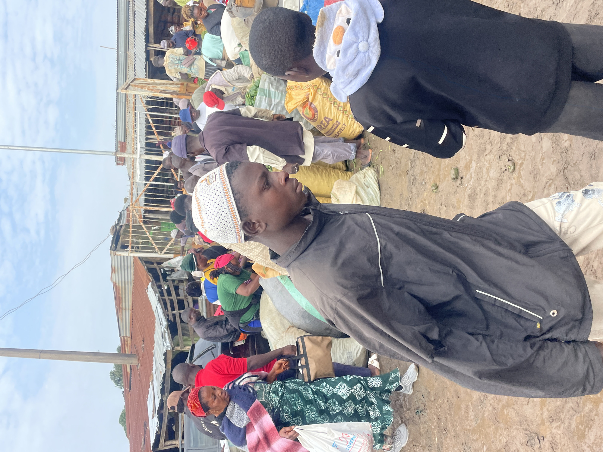 This image captures a busy outdoor market scene in Gura Top, Nigeria. The ground is muddy, suggesting recent rain or the natural state of the market area. In the foreground, two young men are the primary focus. The one on the left, wearing a black jacket and a white and orange patterned skullcap, looks down and to the right. The young man on the right, facing away from the camera, is wearing a black hoodie with white trim and a cartoonish design on the hood.
Behind them, a bustling crowd of people are engaged in market activities. A woman in a green and white patterned dress and a red headscarf is visible on the left, holding a white plastic bag and a brown handbag. She is walking alongside a man in a red shirt. Other individuals are seen amidst piles of sacks, some appearing to contain produce. Some are wearing hats and traditional clothing.
In the background, a row of makeshift market stalls with corrugated iron roofs are present, indicating a semi-permanent market structure. A tall pole, possibly a utility pole, stands prominently in the sky. The overall atmosphere is one of lively commerce in a somewhat rustic and unpaved environment.